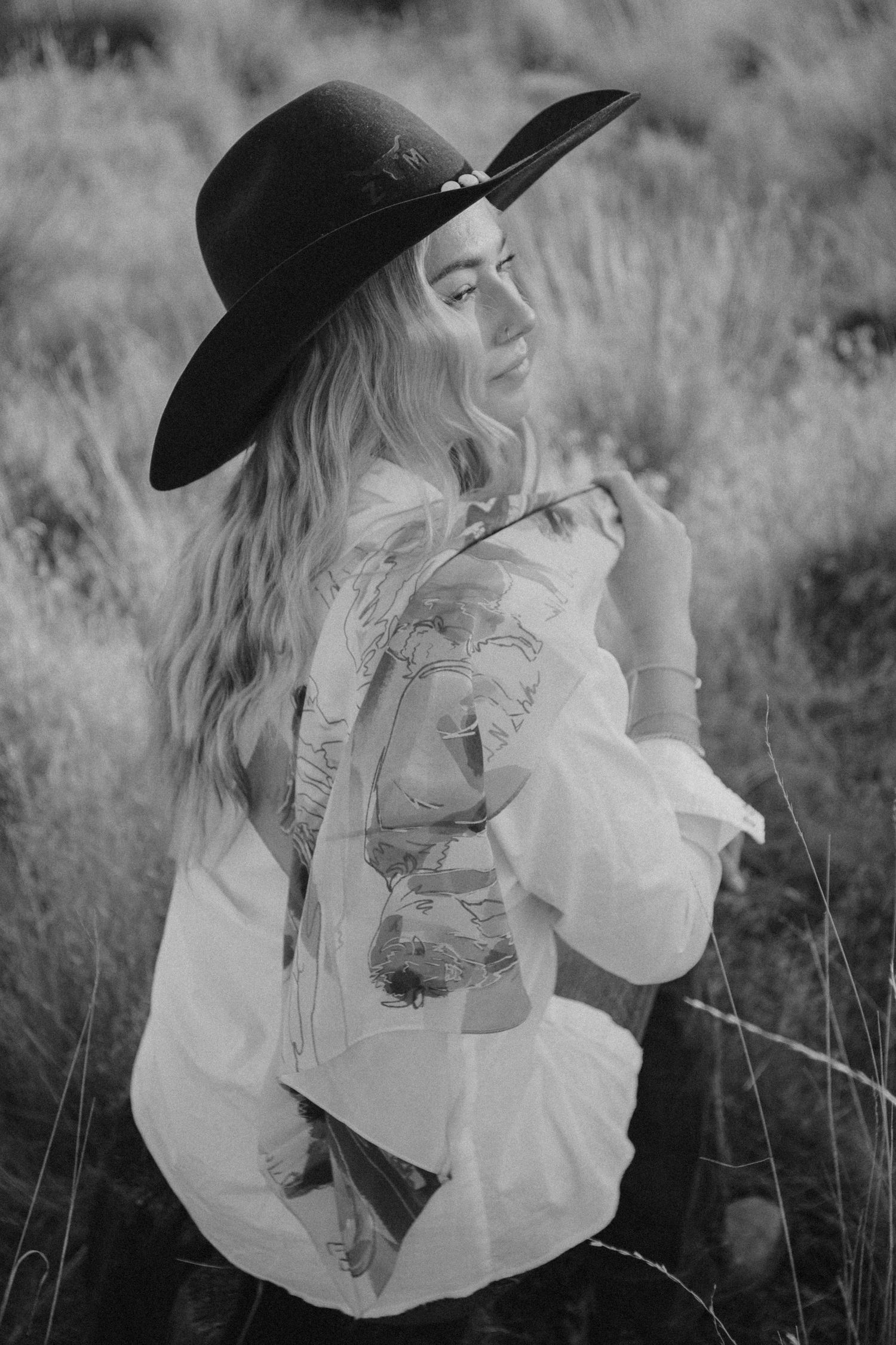 Person in black hat and white ruffled blouse poses dramatically in black and white portrait.