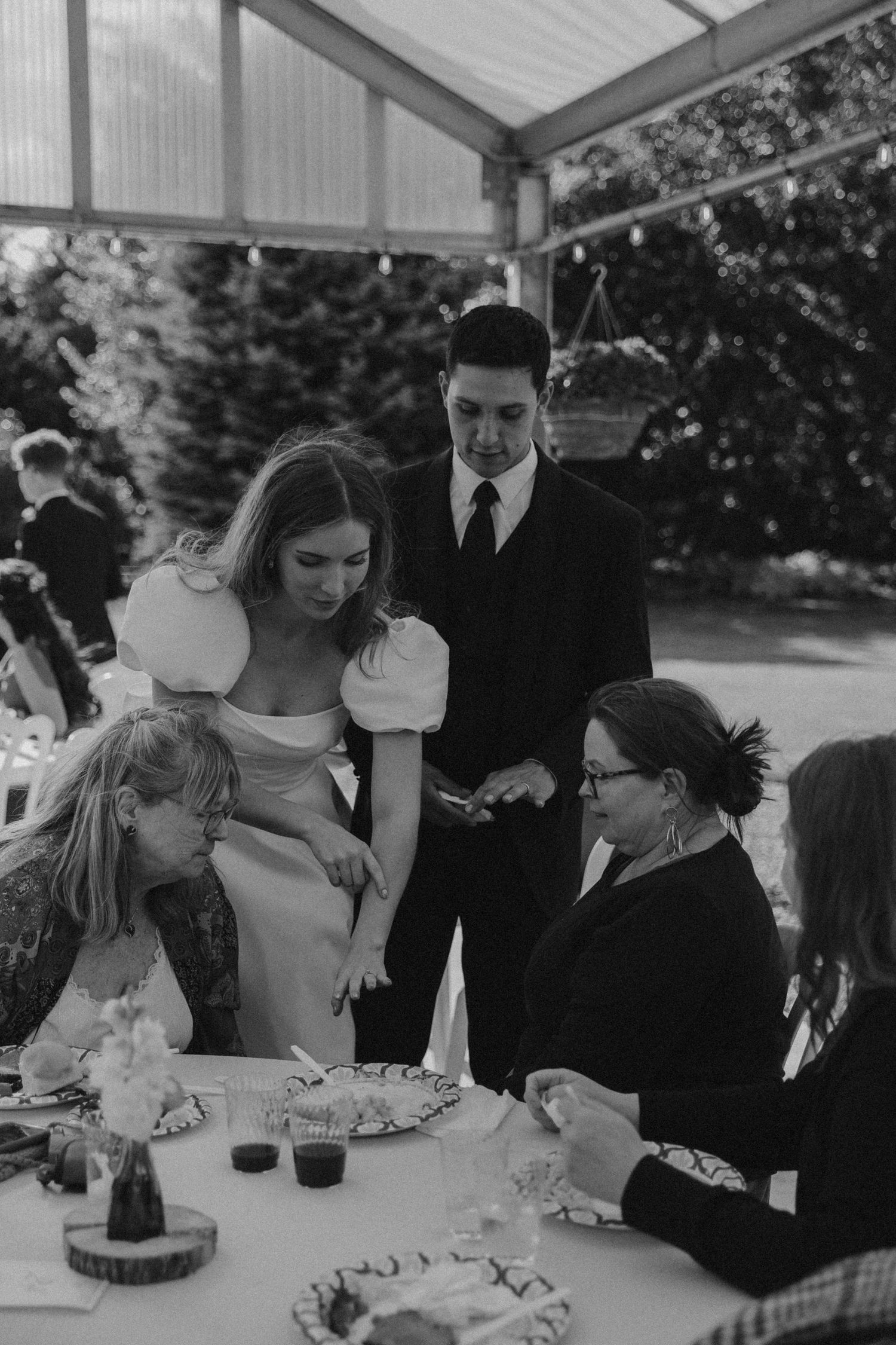Wedding guests gather around tables under a covered pavilion during the reception celebration.