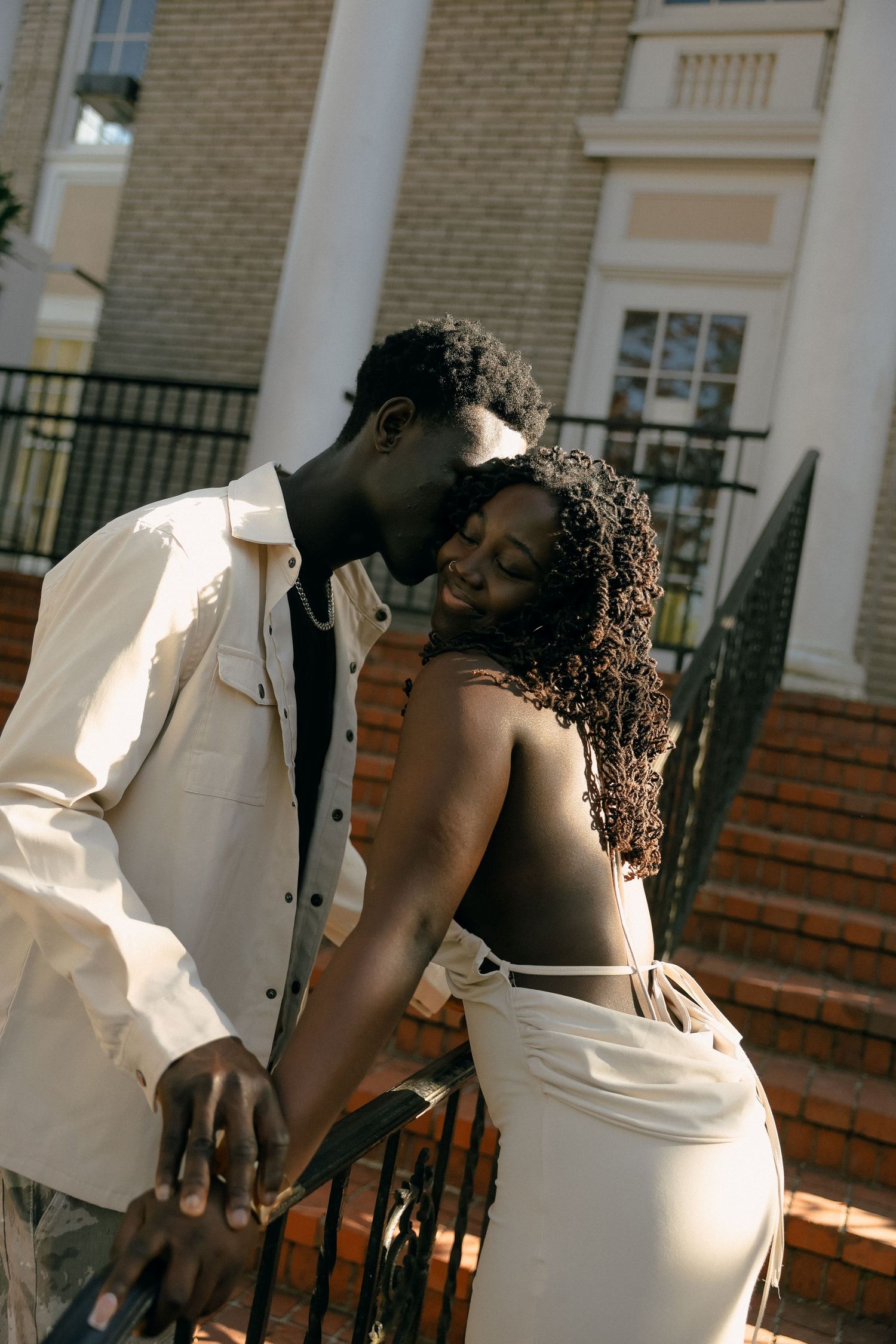 A couple in elegant white outfits share a tender moment on brick steps.