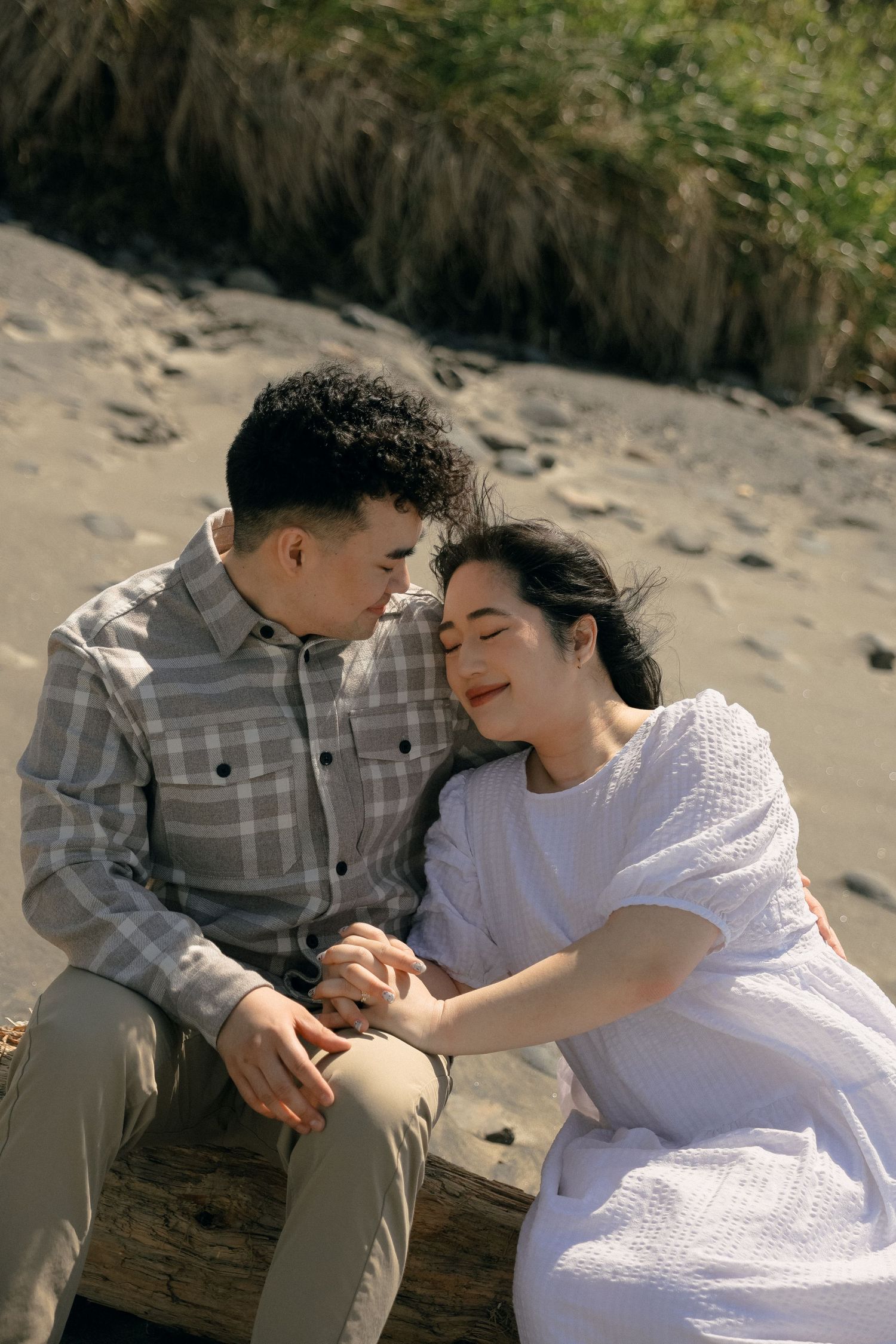 A couple share tender moments while sitting on a beach, dressed in casual attire.