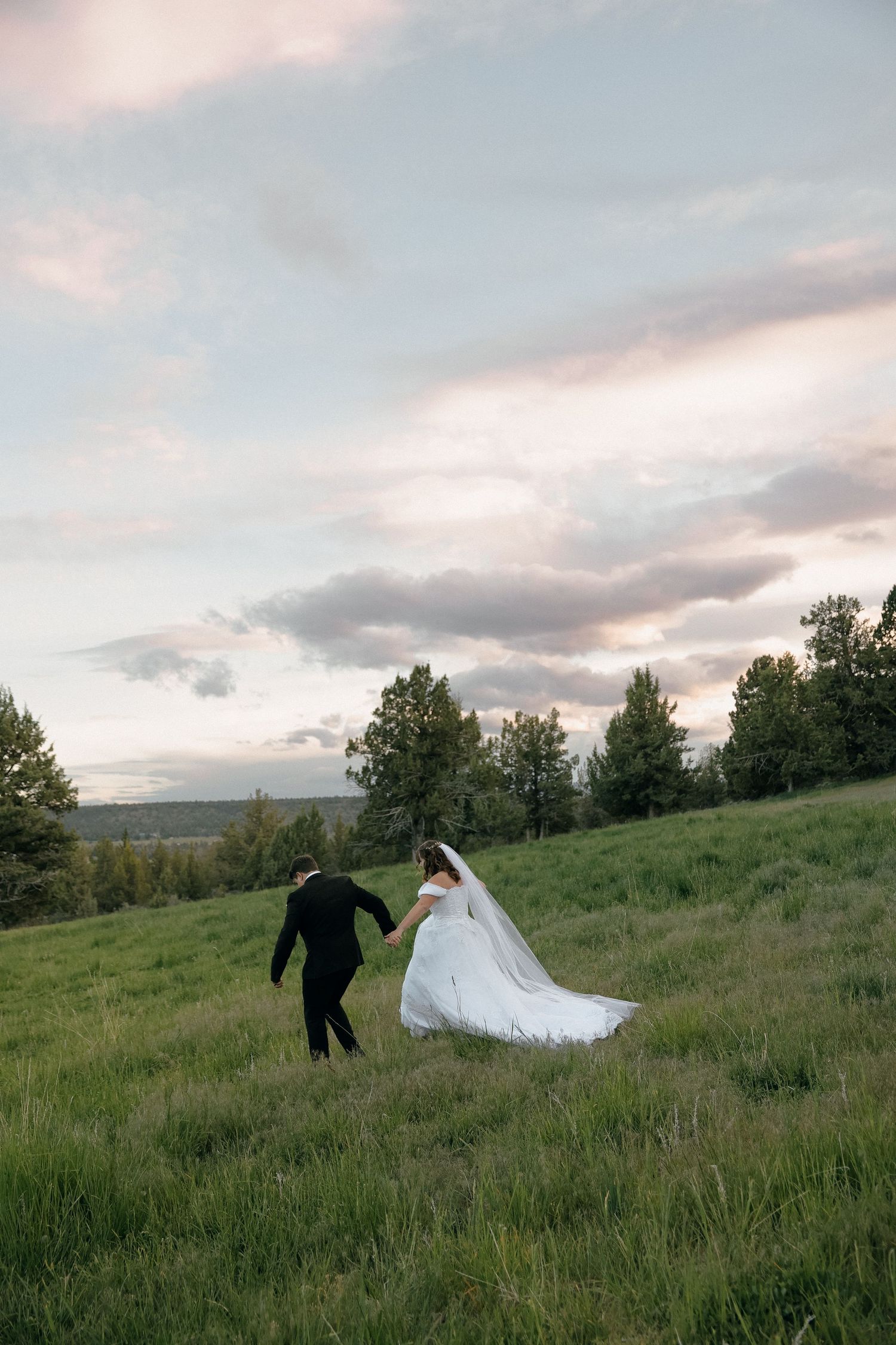 A scenic wedding photo of a couple walking through a grassy hillside at sunset.