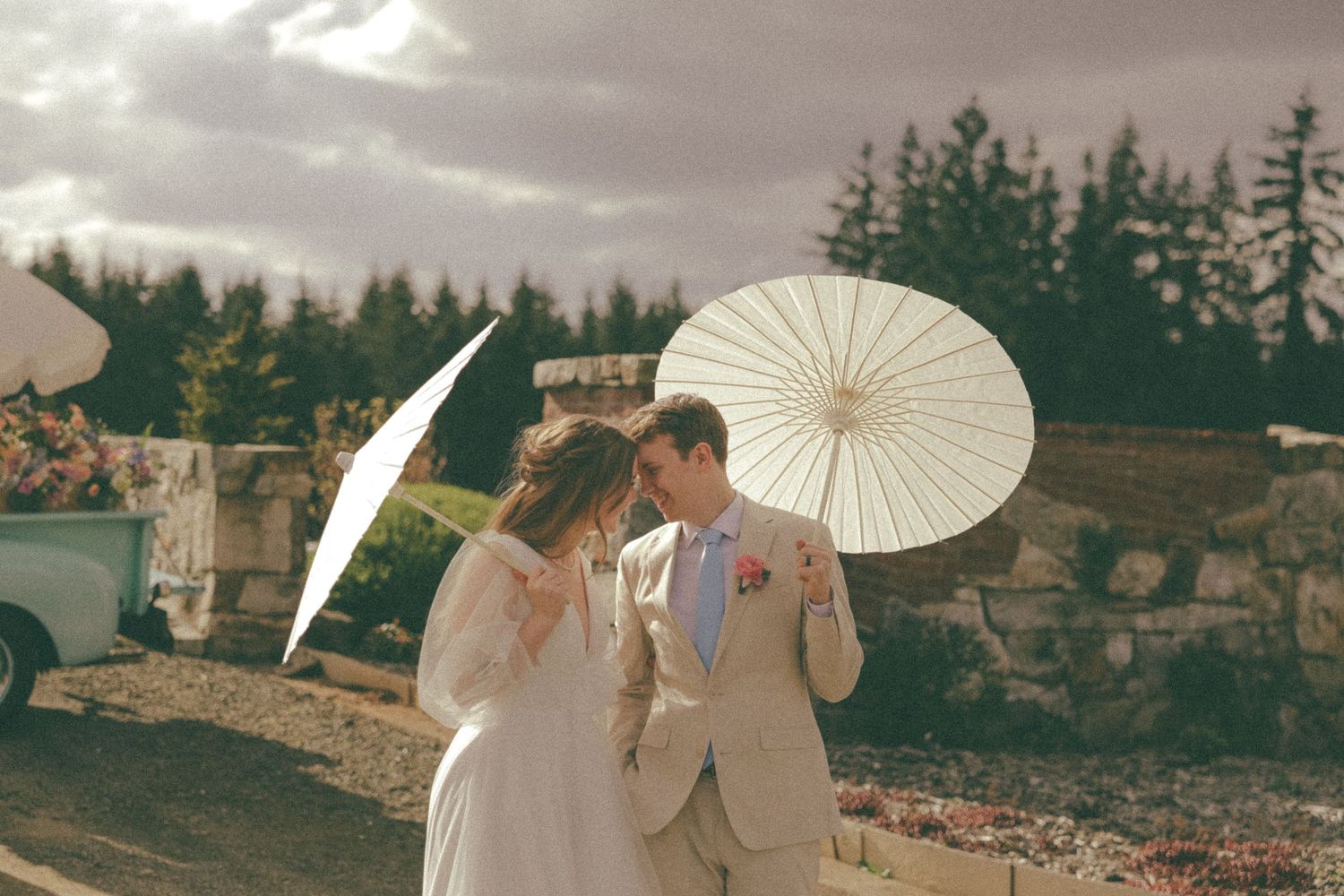 Wedding couple holding white parasols during outdoor ceremony with evergreen backdrop.