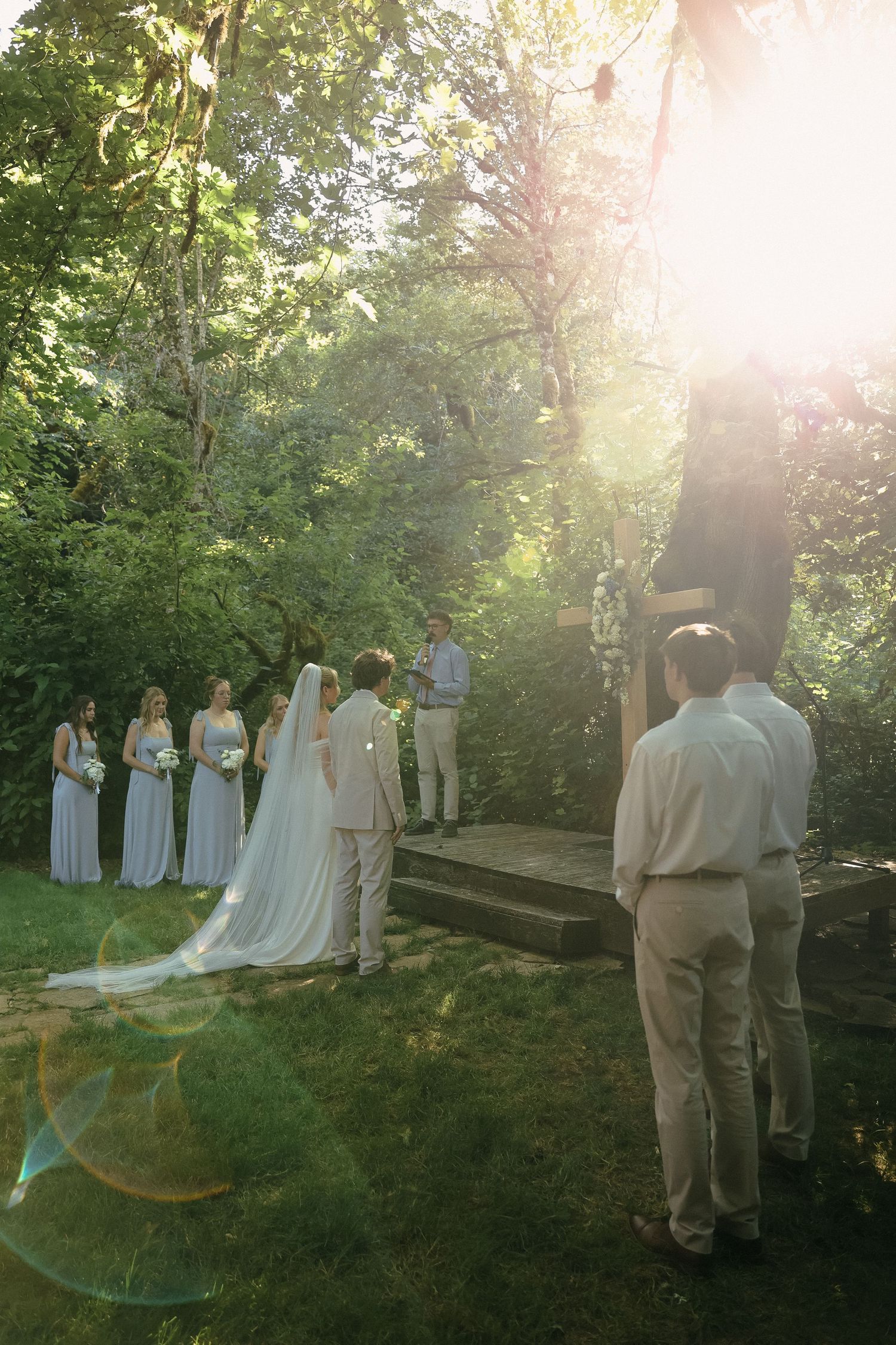 A wedding ceremony takes place in a sunlit forest clearing with attendants in light-colored dresses and suits.