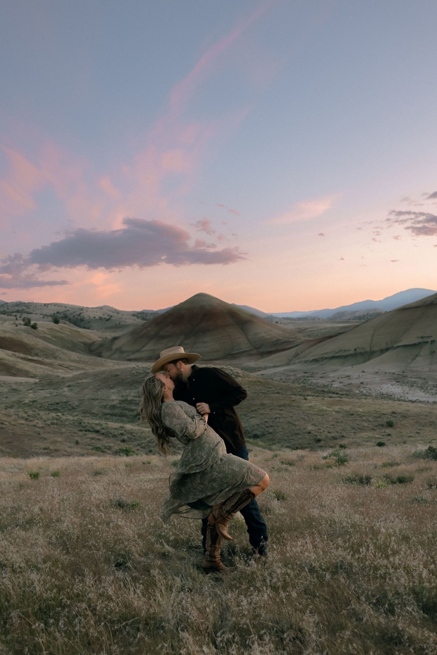 A couple embraces in a romantic dance pose against a backdrop of rolling hills and pink sunset sky.