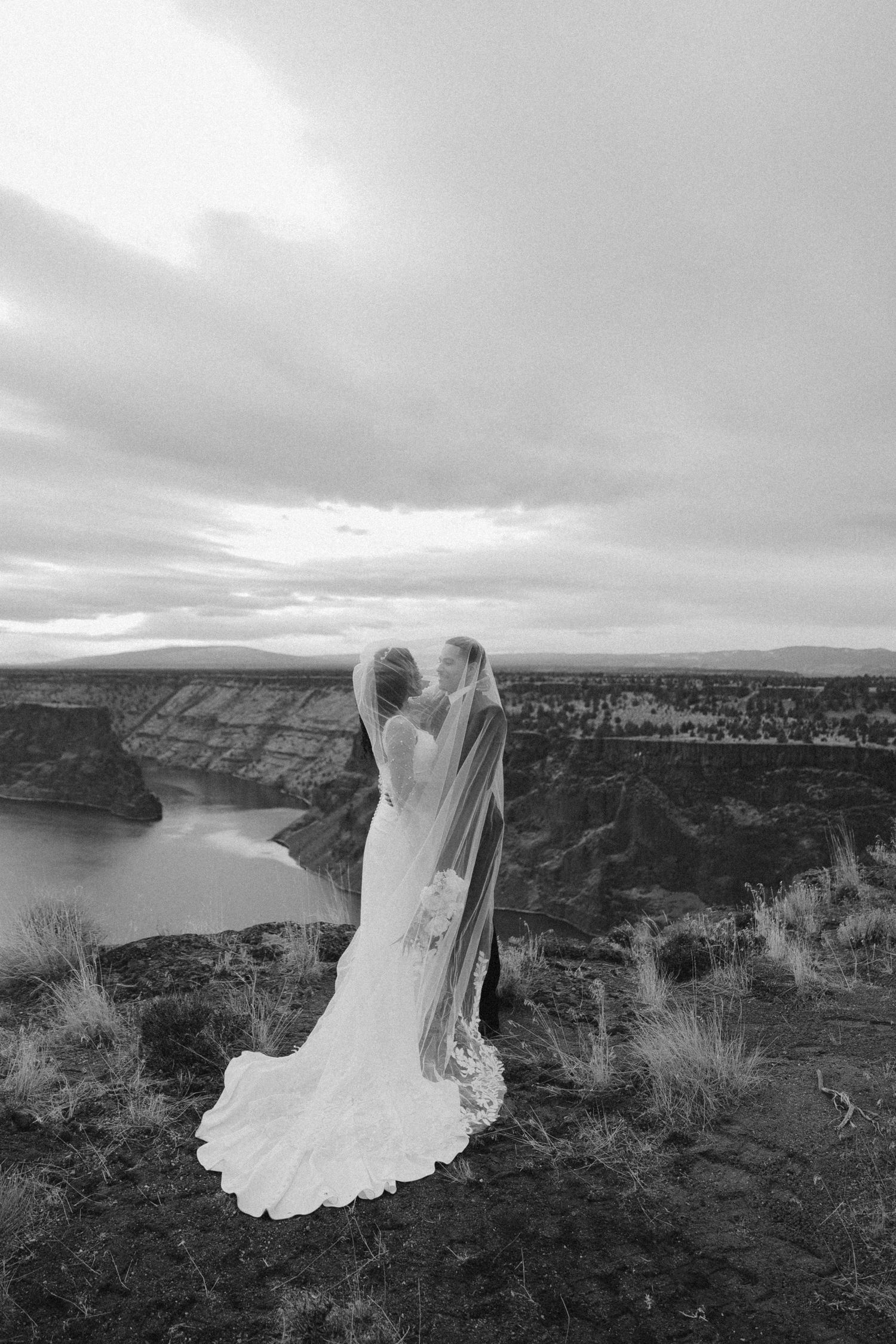 A bride in a flowing white dress and veil stands at the edge of a dramatic canyon overlook in black and white.