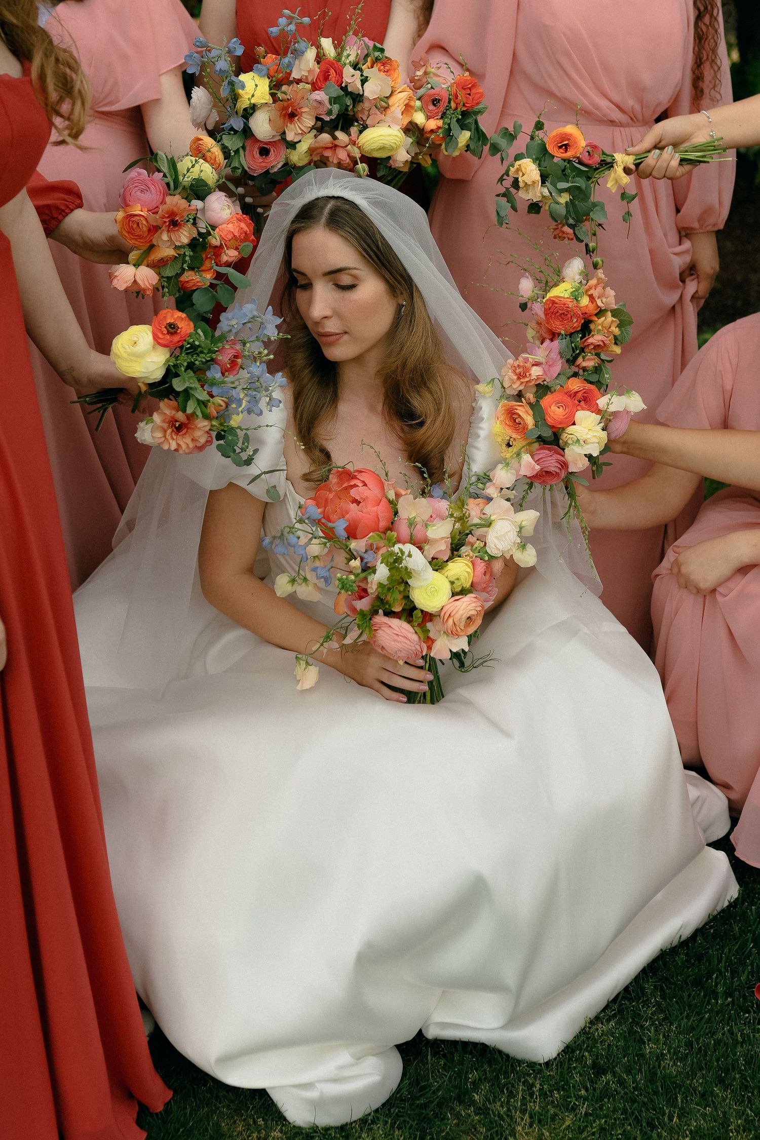 A bridal portrait featuring a white gown, floral arch, and coral bridesmaid dresses in an outdoor setting.