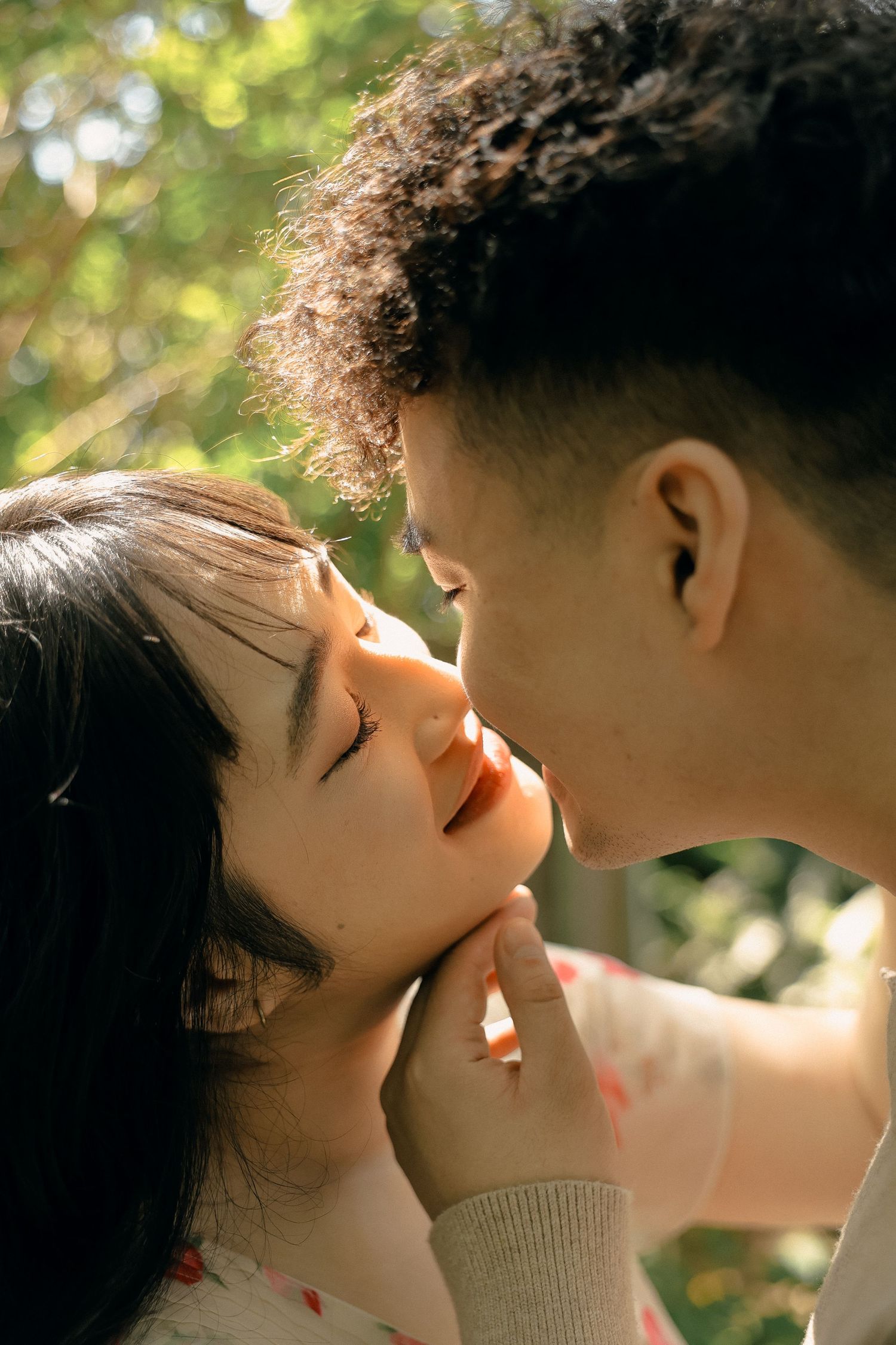 A couple shares a tender kiss in dappled sunlight filtering through trees.