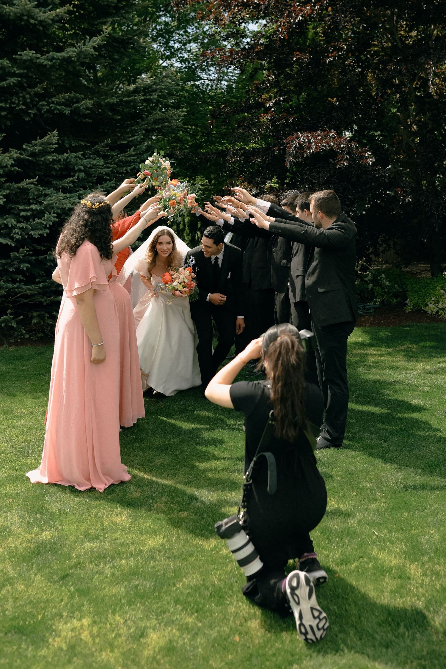 Wedding party in pink dresses and black suits poses for creative photos on a lawn.
