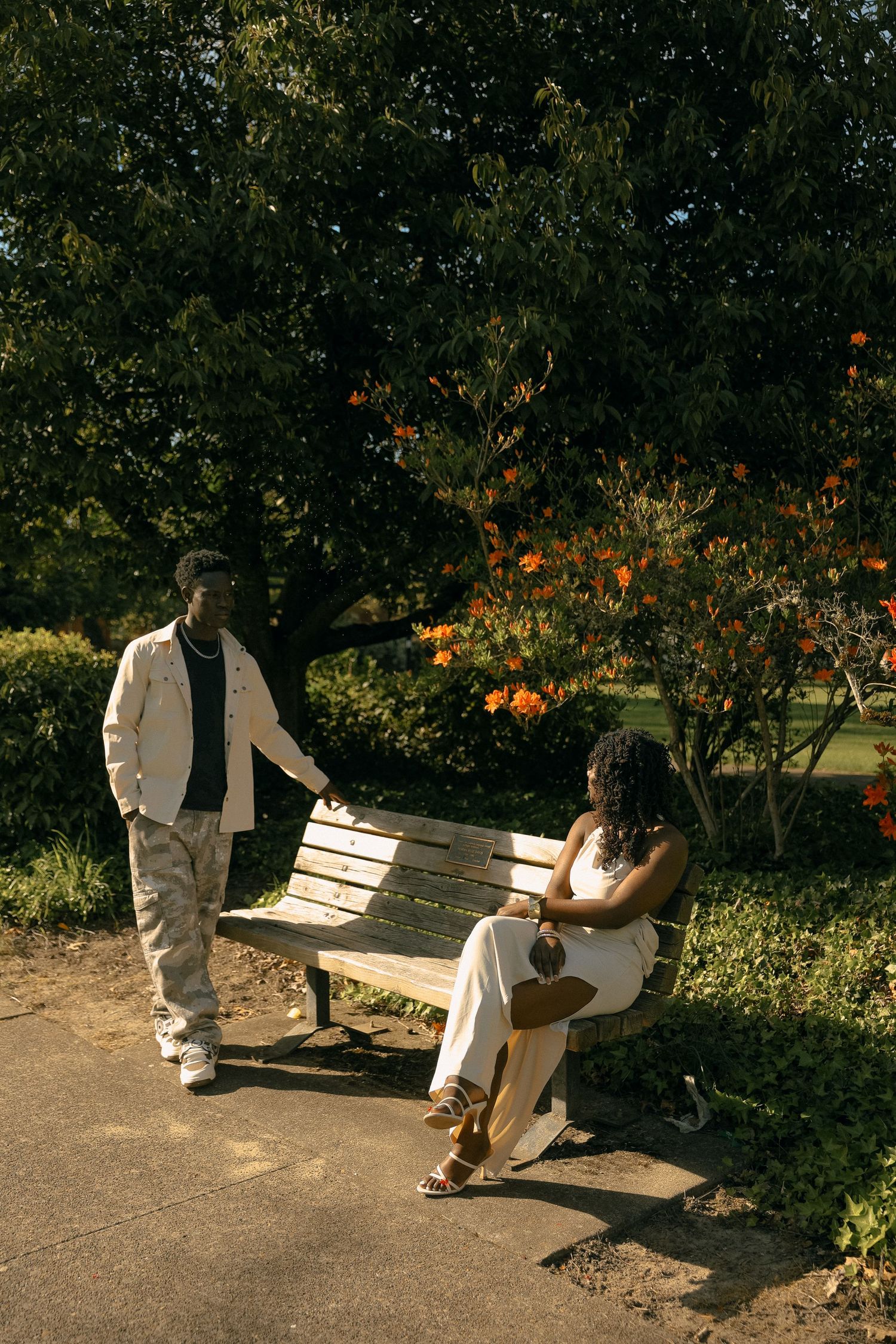 A couple shares a moment near a park bench surrounded by flowering trees in sunlight.