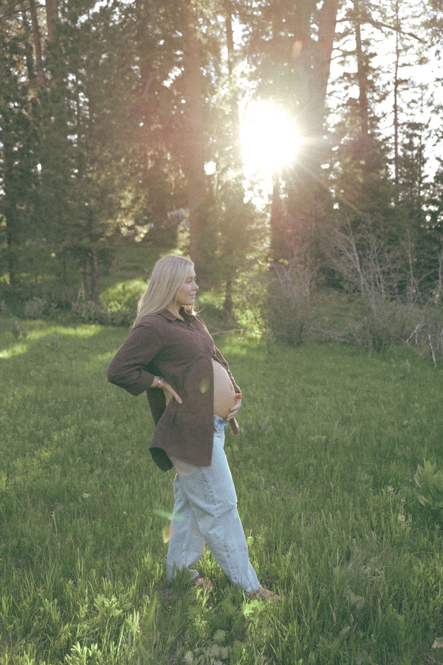 A pregnant person stands in a sunlit meadow surrounded by evergreen trees at golden hour.