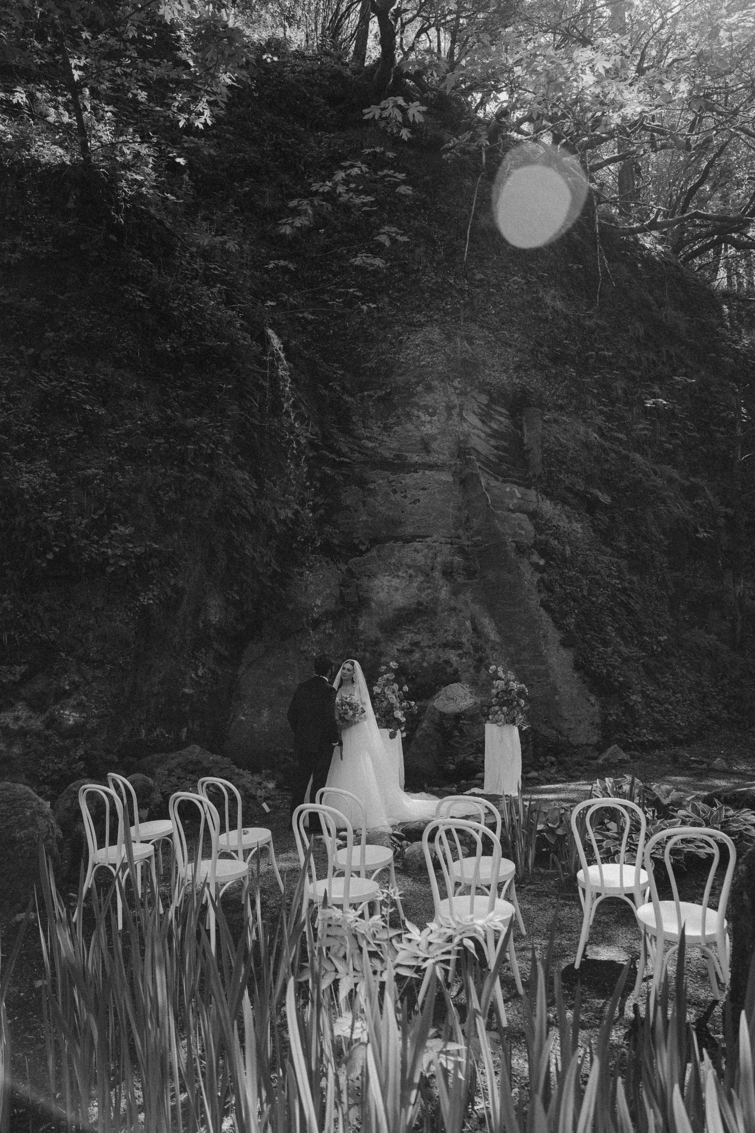 Empty white chairs arranged outdoors for wedding ceremony with natural wooded backdrop.