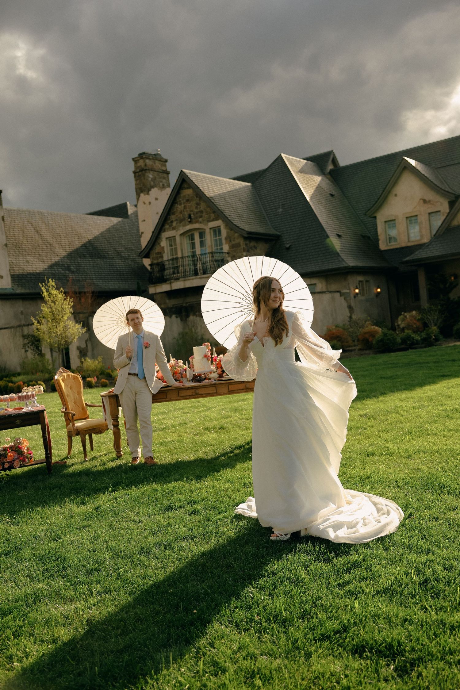 A bride with a white parasol poses in front of a grand estate under stormy skies.