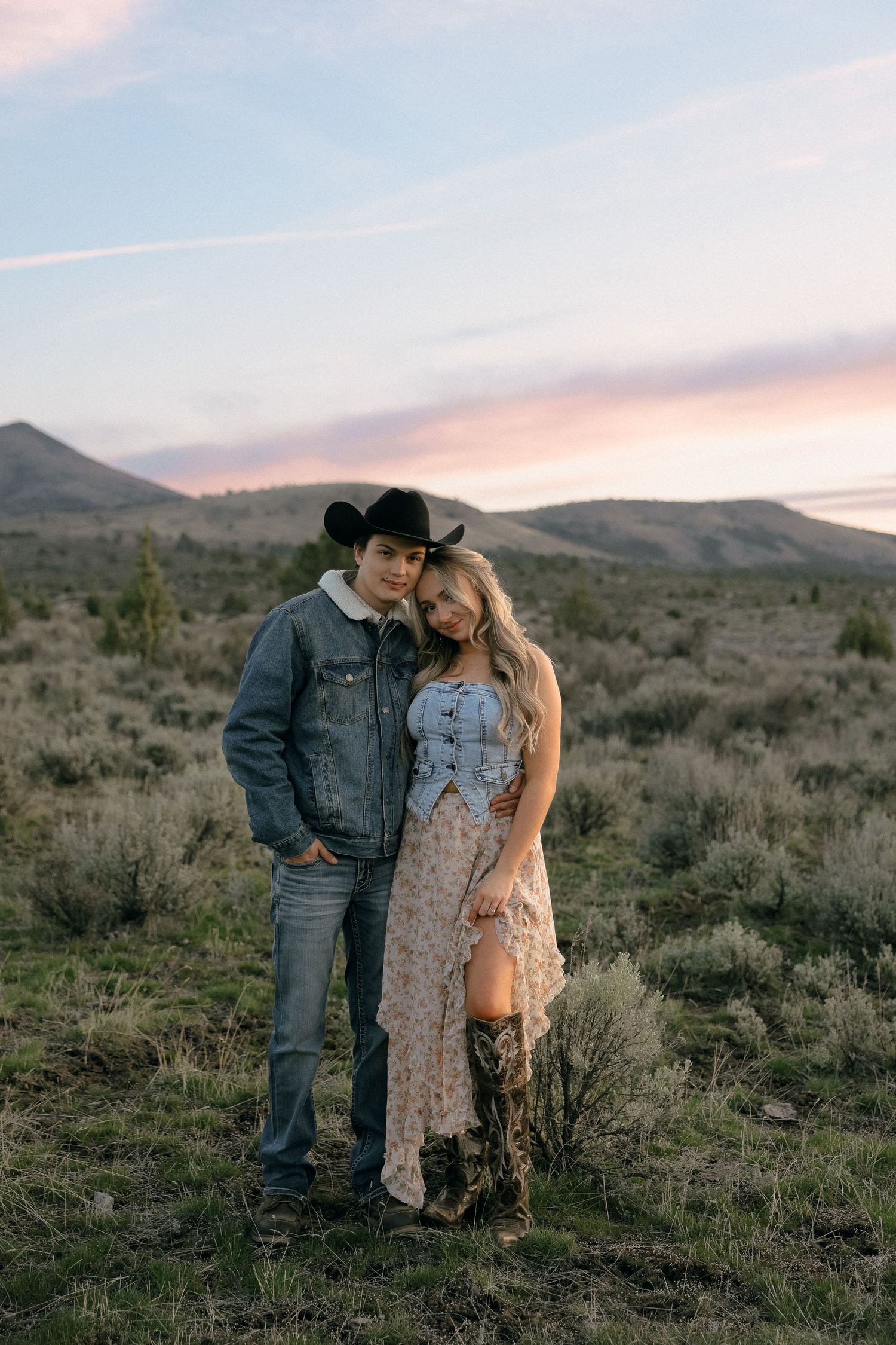 A couple in western wear embraces against a desert mountain backdrop at sunset.