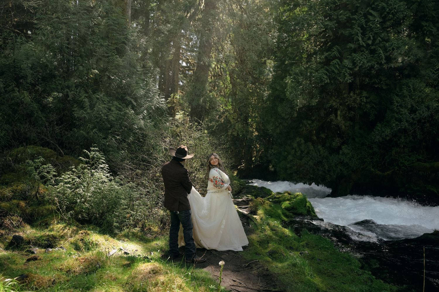 A couple in wedding attire stands on a forest path beside a rushing stream in dappled sunlight.
