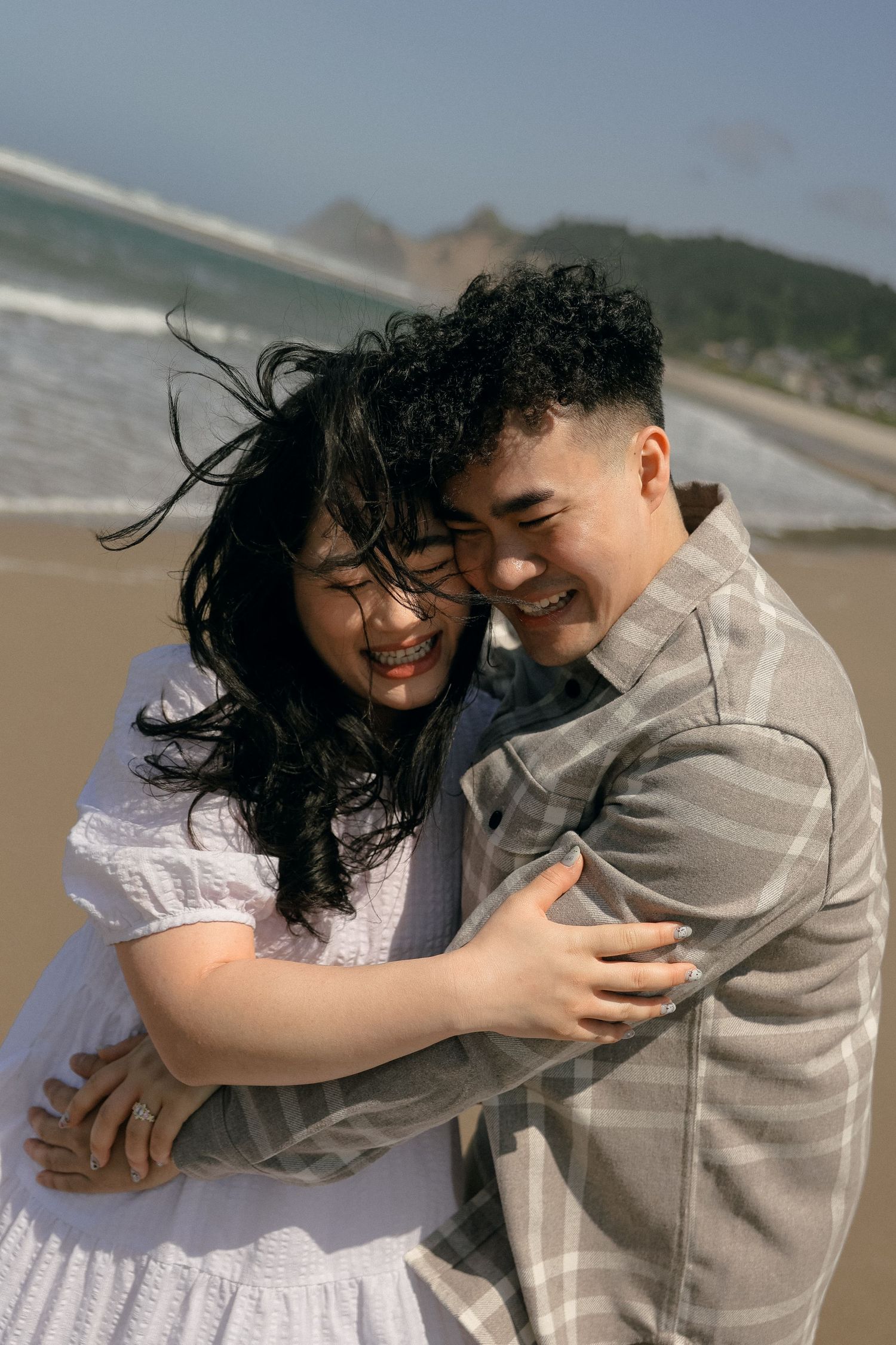 A couple embracing and laughing together on a sandy beach with mountains in the distance.