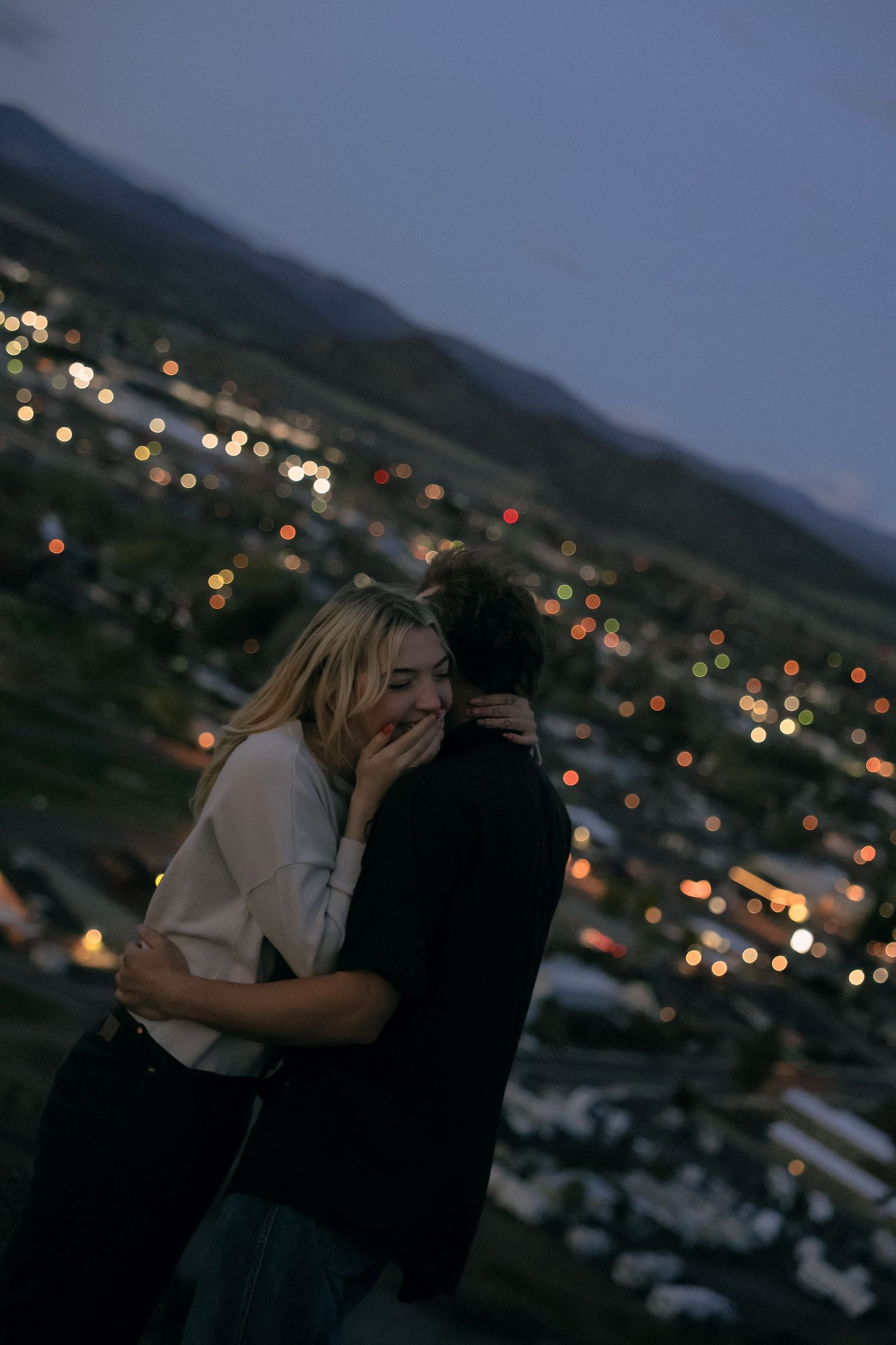 A couple shares an intimate moment overlooking city lights on a hillside at dusk.