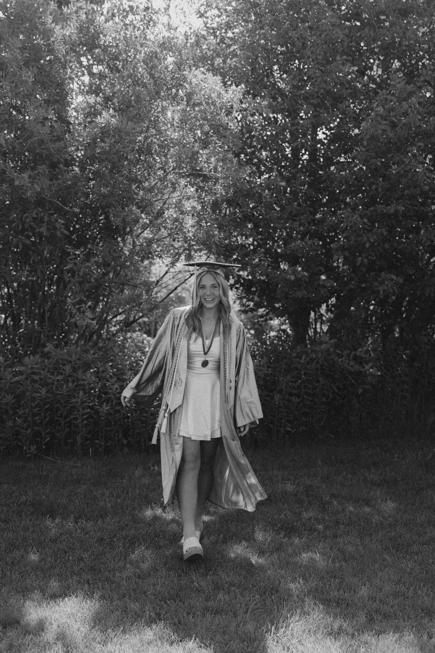 Graduate in flowing graduation gown dancing joyfully in a grassy outdoor setting.
