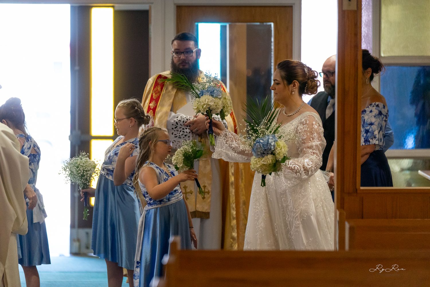 Wedding party members gathered during ceremony with blue bridesmaid dresses and floral bouquets.