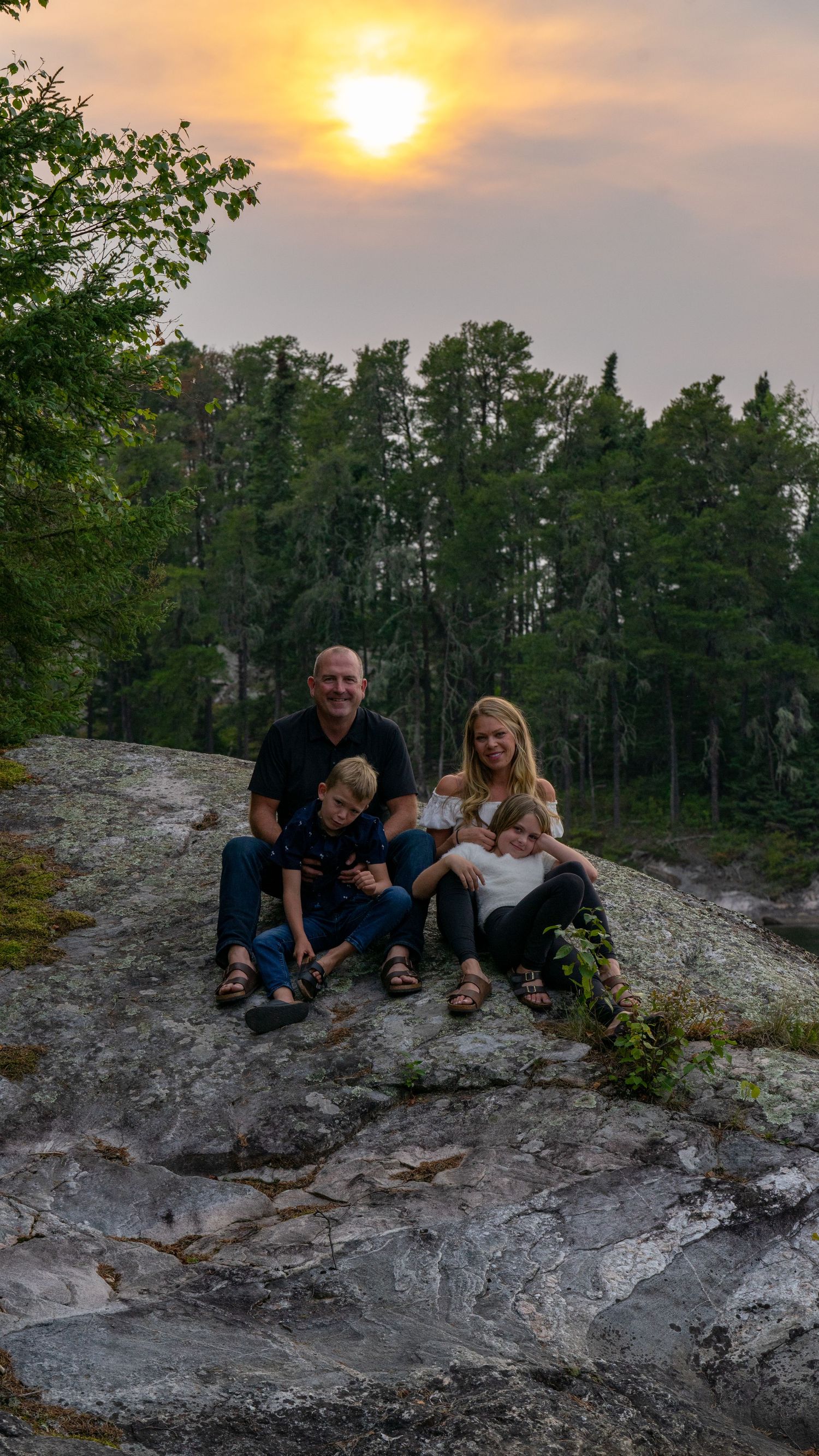 Family portrait on rocky cliff during golden sunset with evergreen forest background.