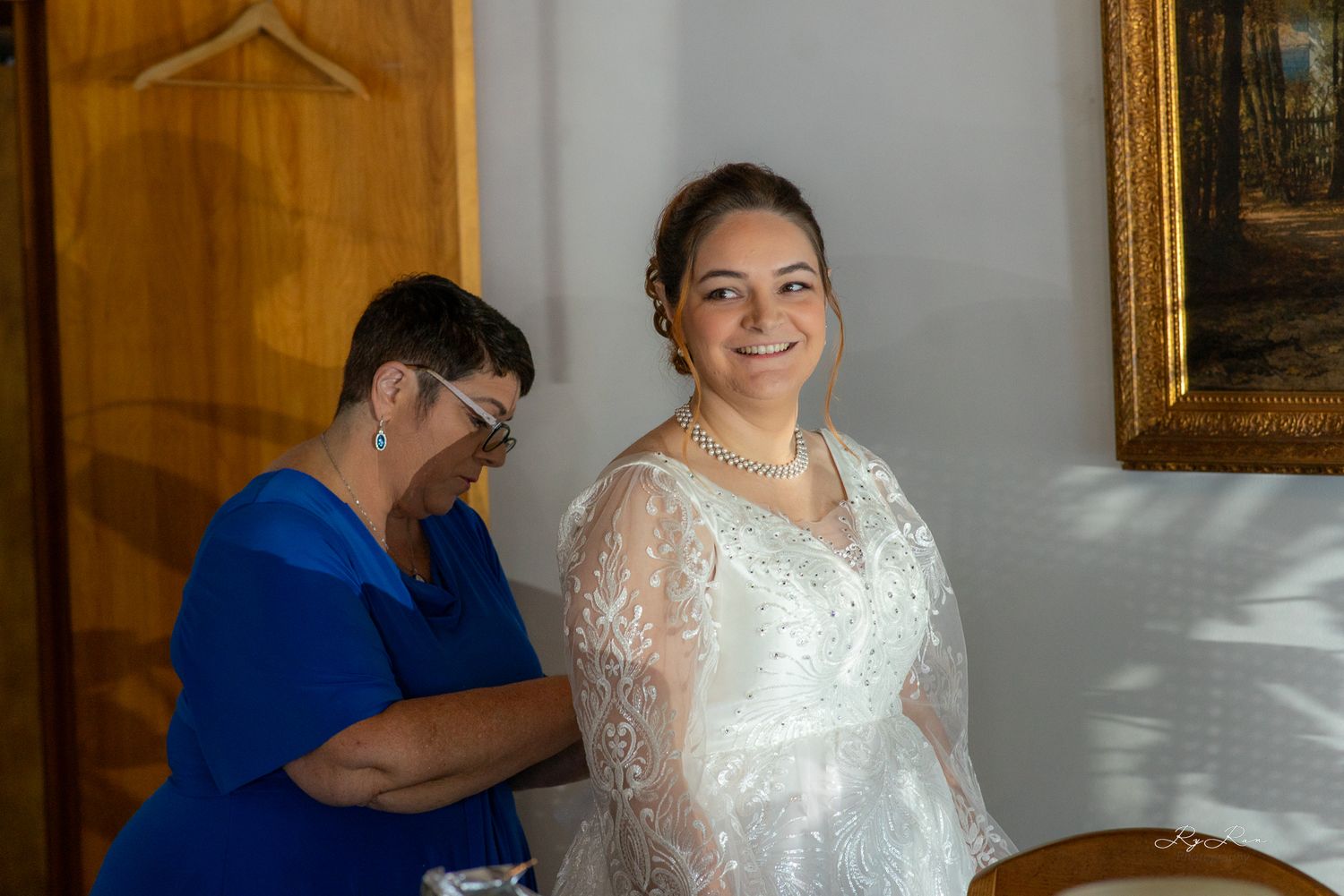 A bride in white lace wedding dress stands while getting final adjustments.
