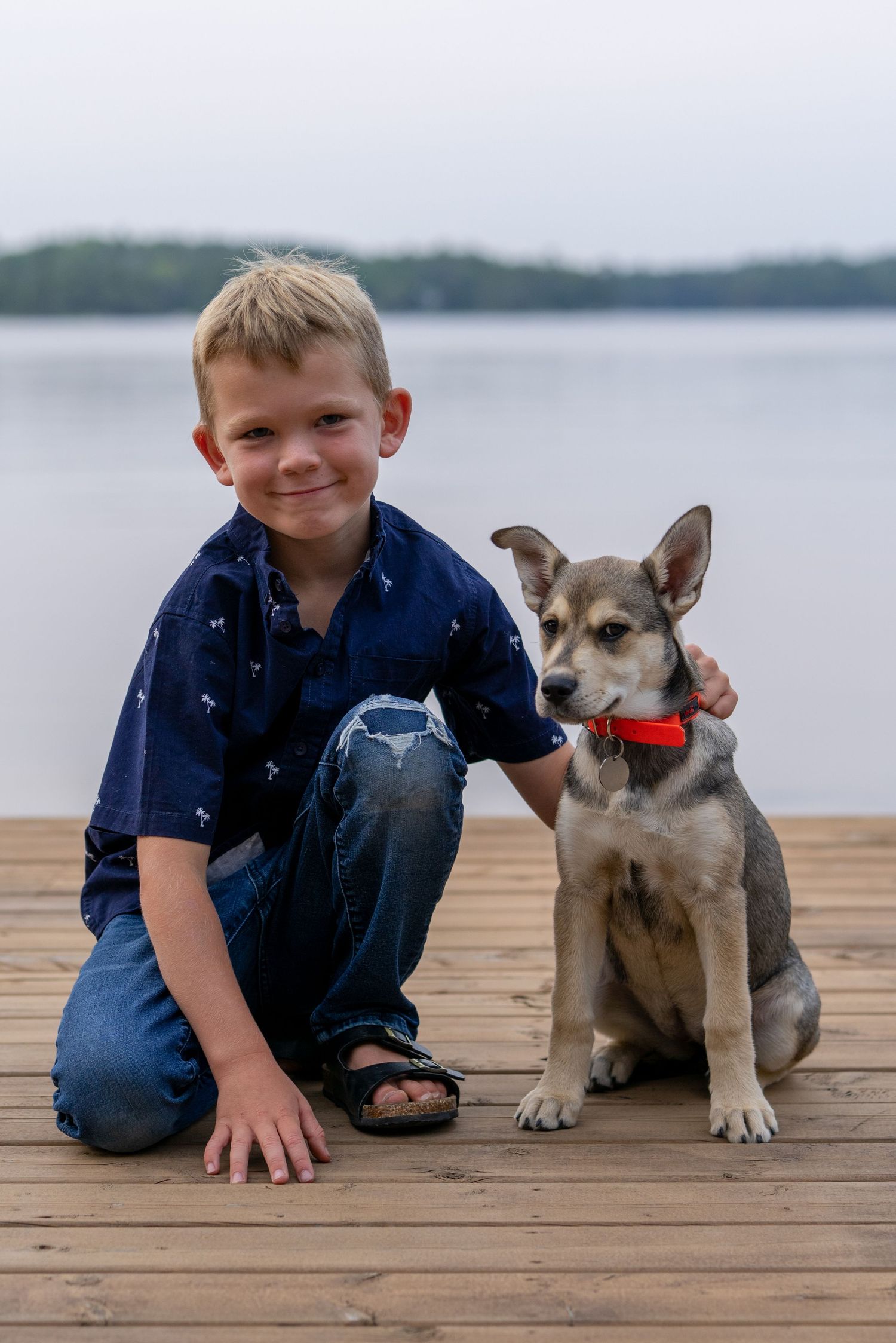 A young friend poses with their loyal dog wearing an orange collar on a lakeside dock.