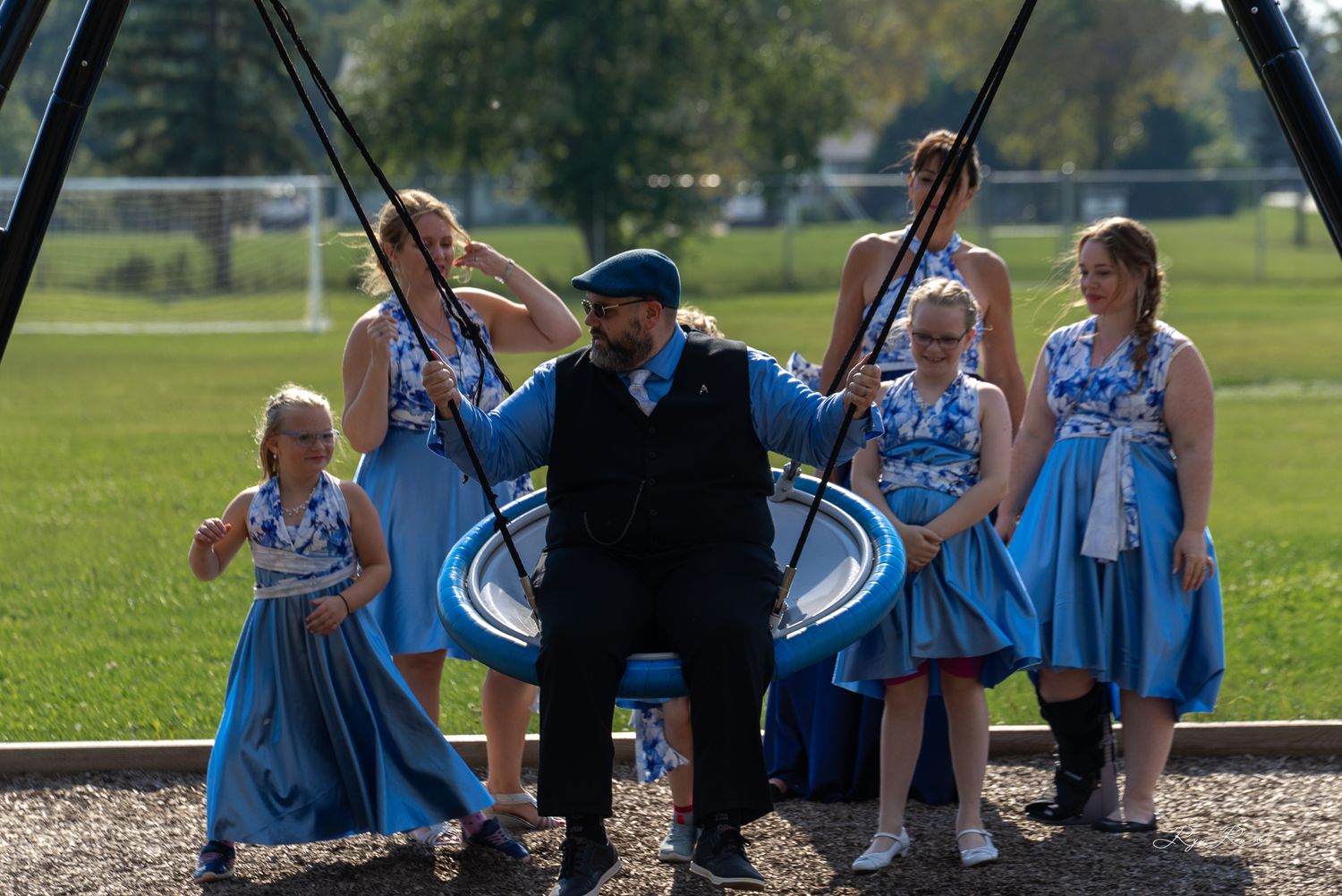Wedding party enjoying a playful moment on outdoor playground equipment in blue dresses and formal attire.