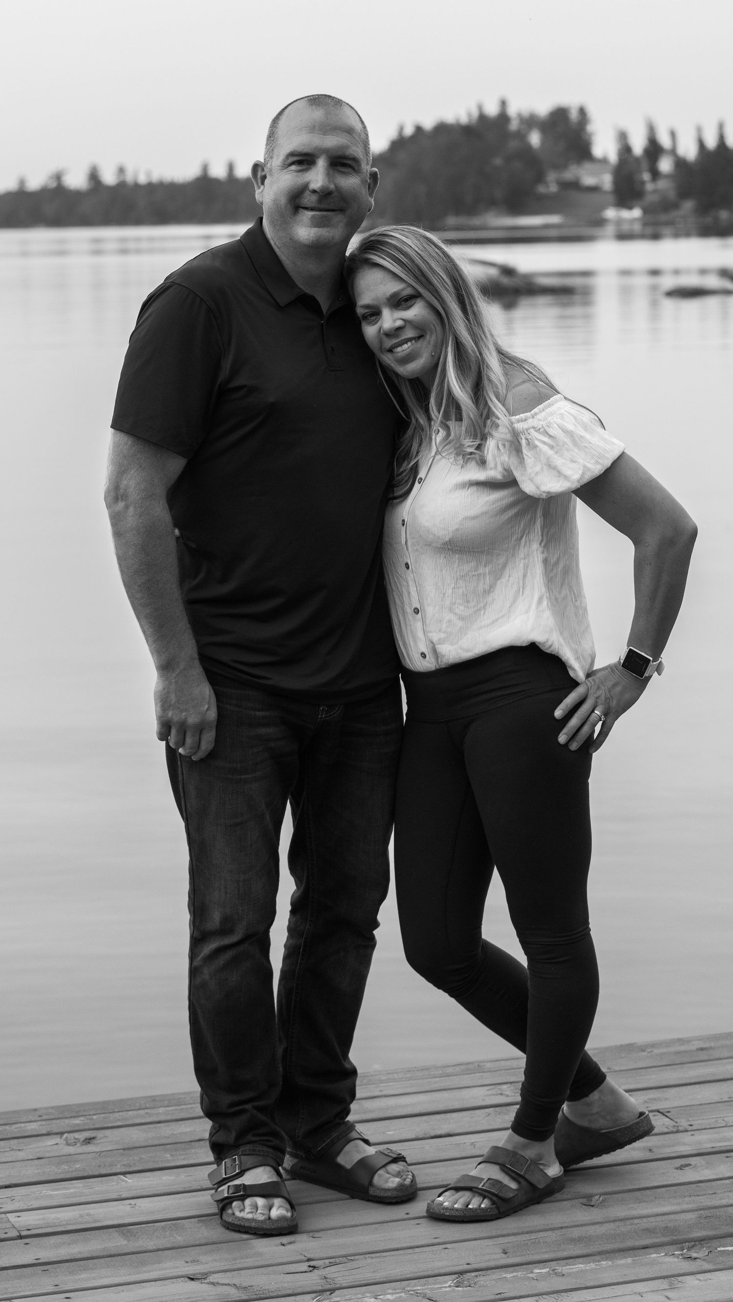 A black and white portrait of a couple standing close together on a lakeside dock.