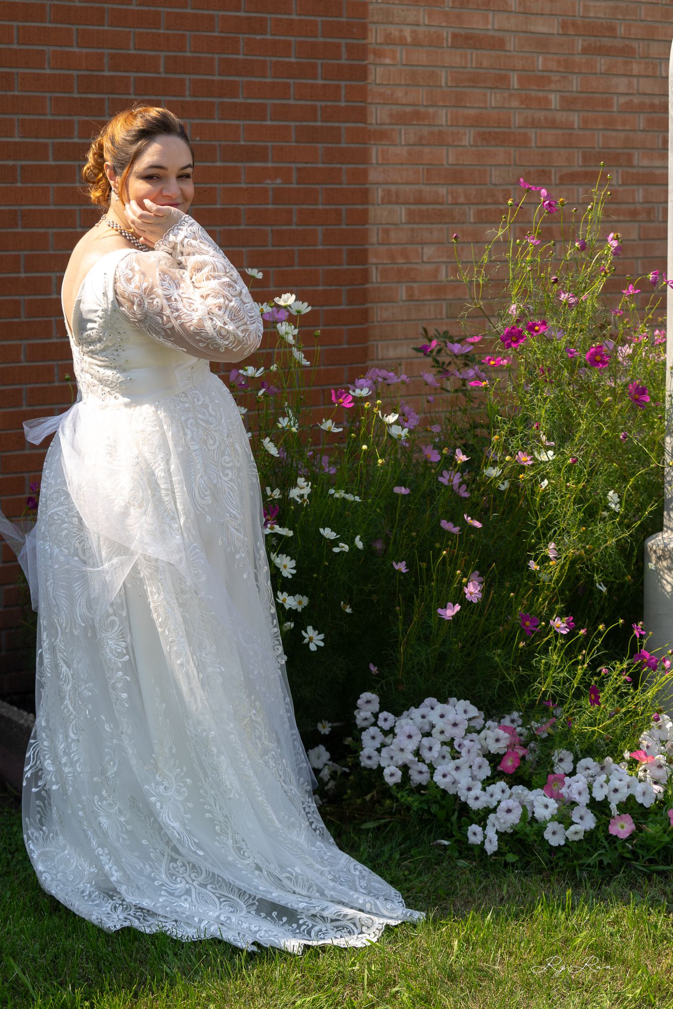 Bride in elegant lace wedding gown poses by garden with colorful flowers.