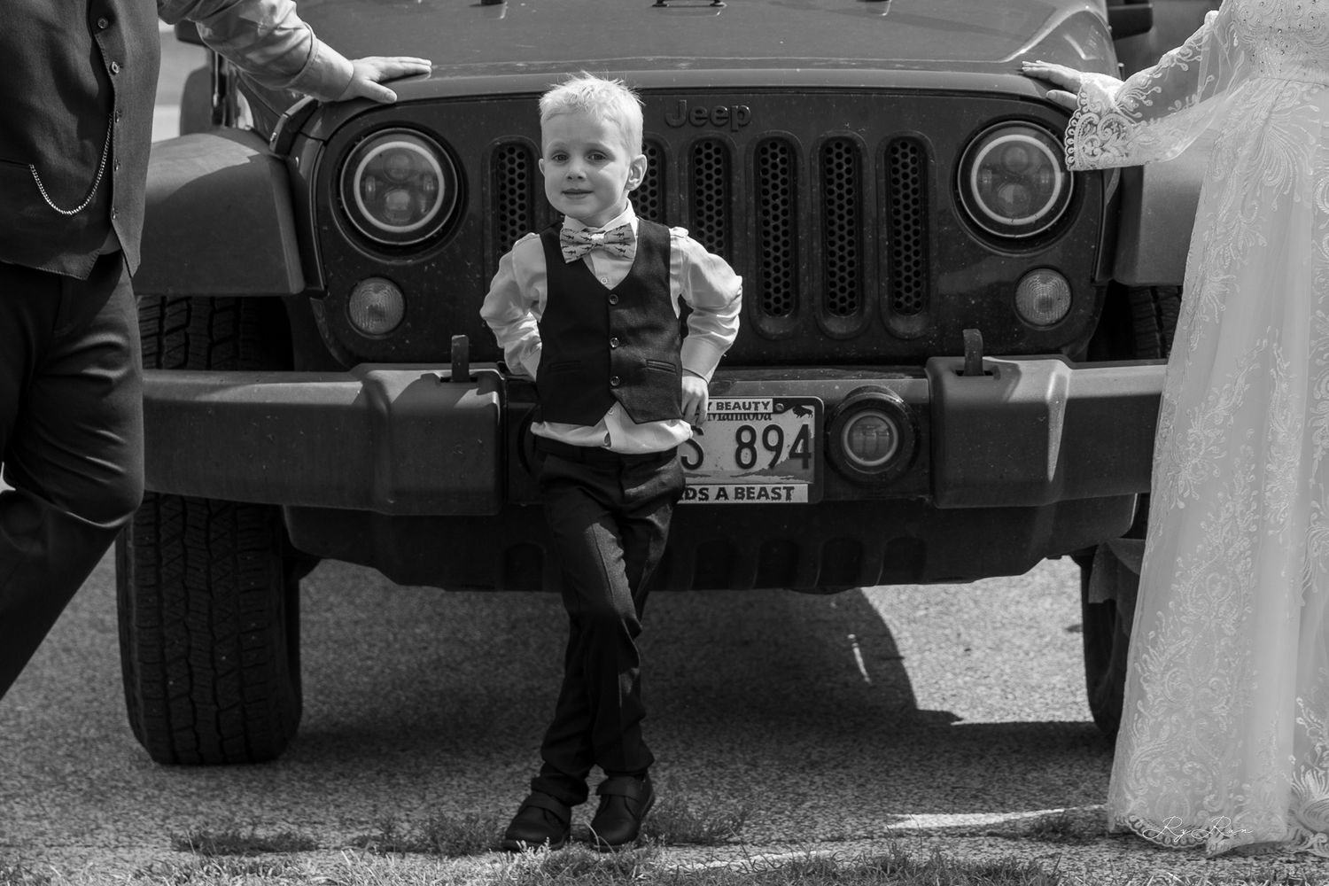 A black and white photo of a young attendant posing in front of a Jeep grill at a wedding.