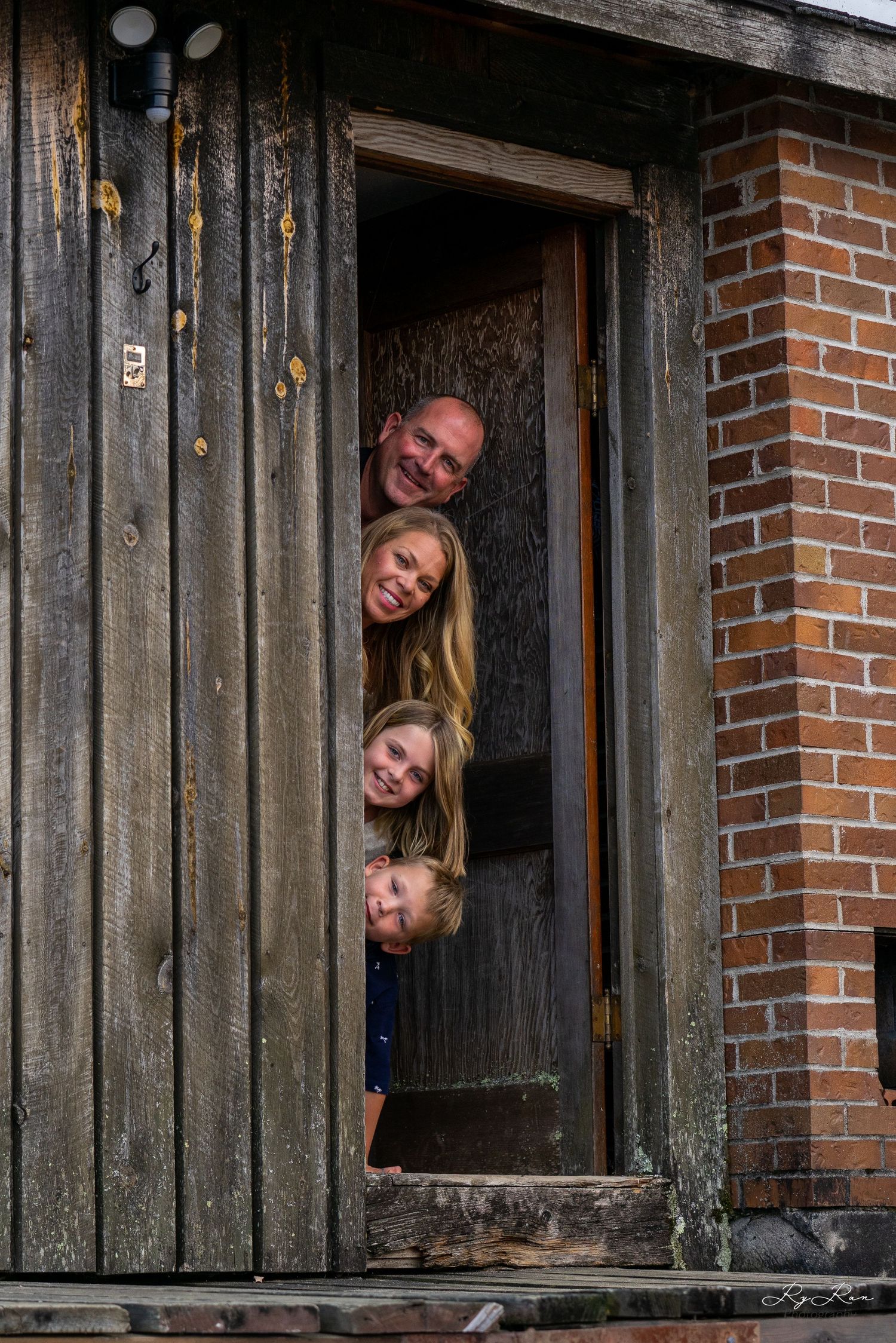 Family members playfully stacked in doorway of weathered wooden building.