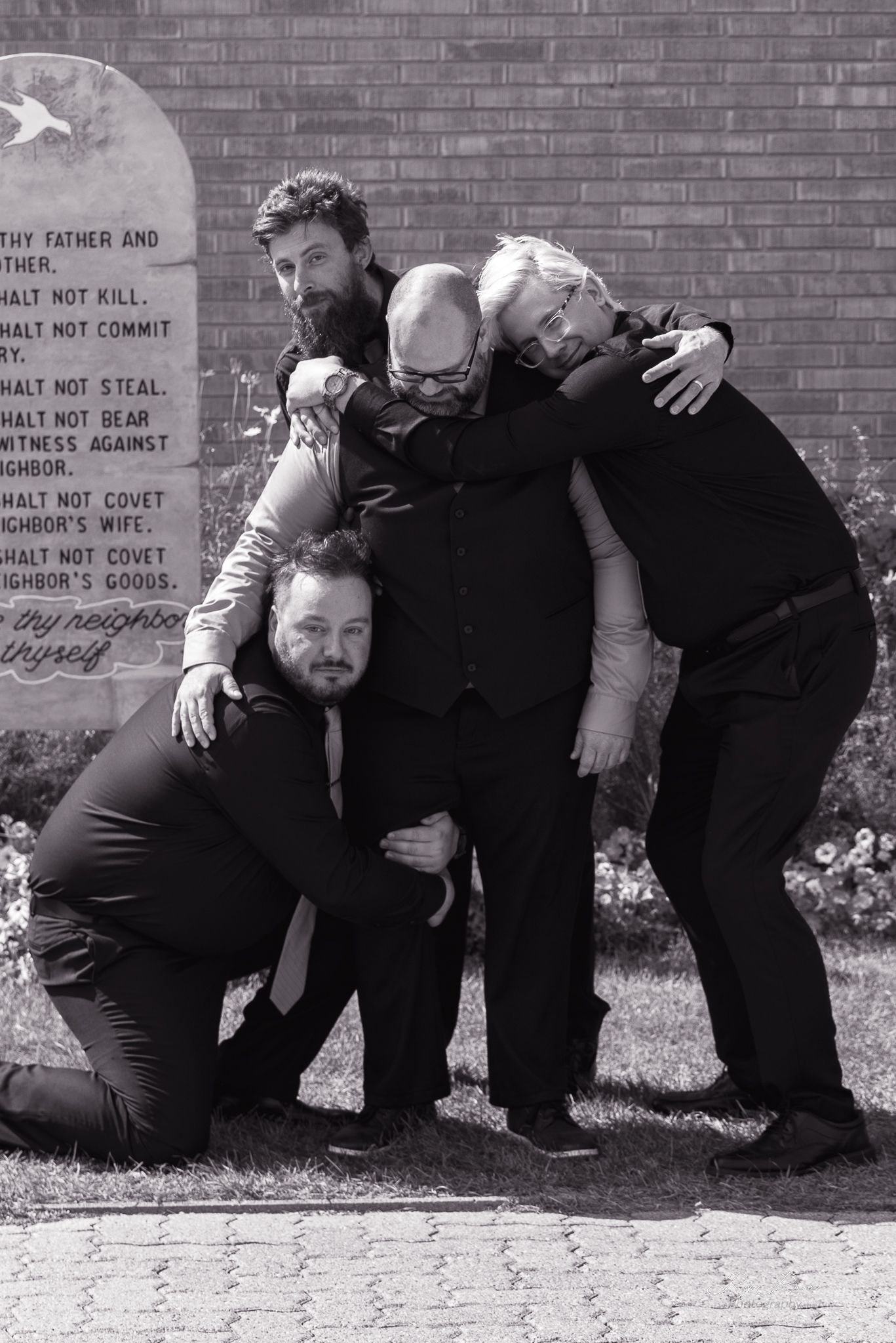 Four friends embrace in emotional group hug against brick wall with stone tablets.