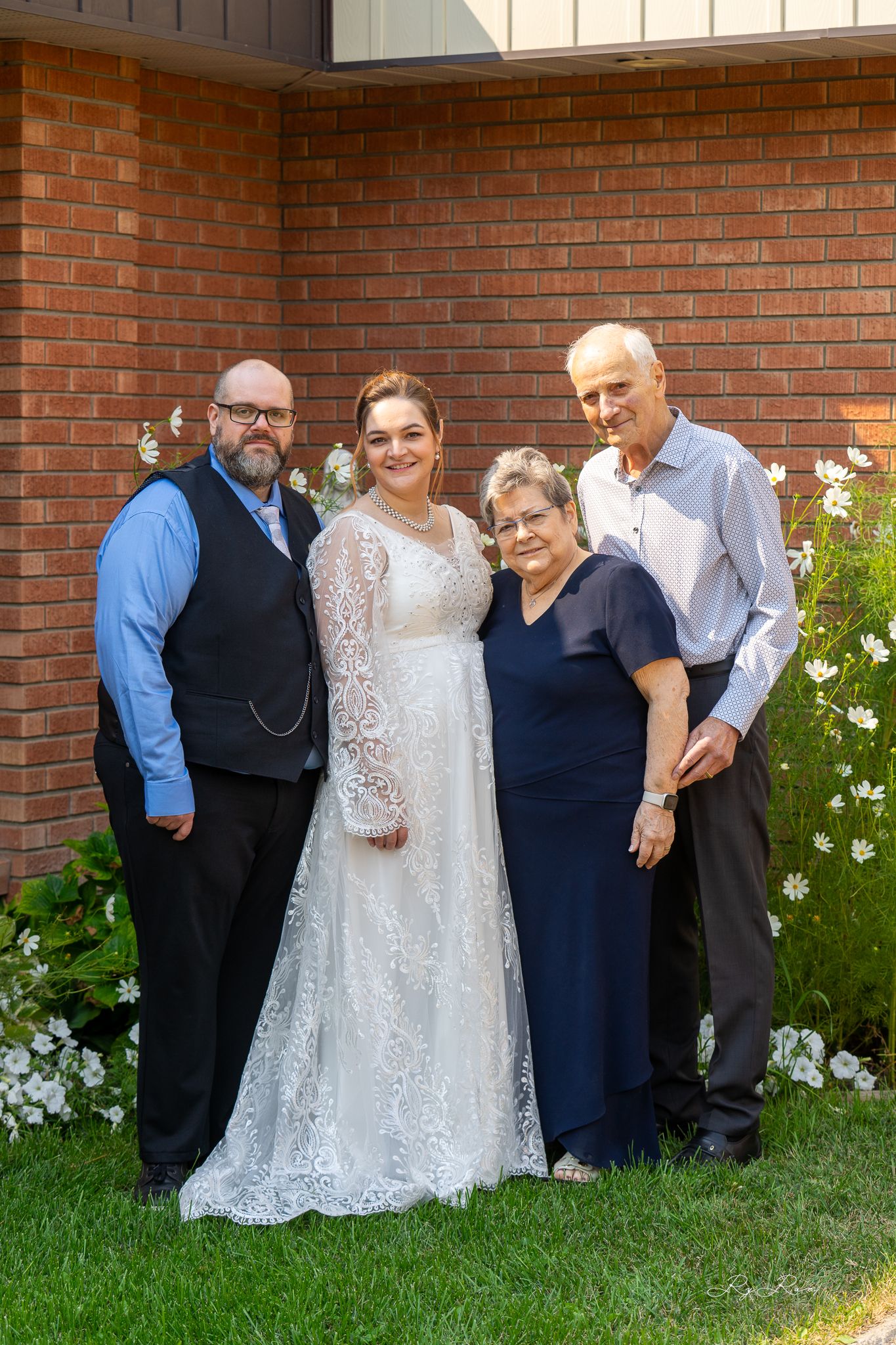 Wedding day family portrait against brick wall with blooming garden flowers.