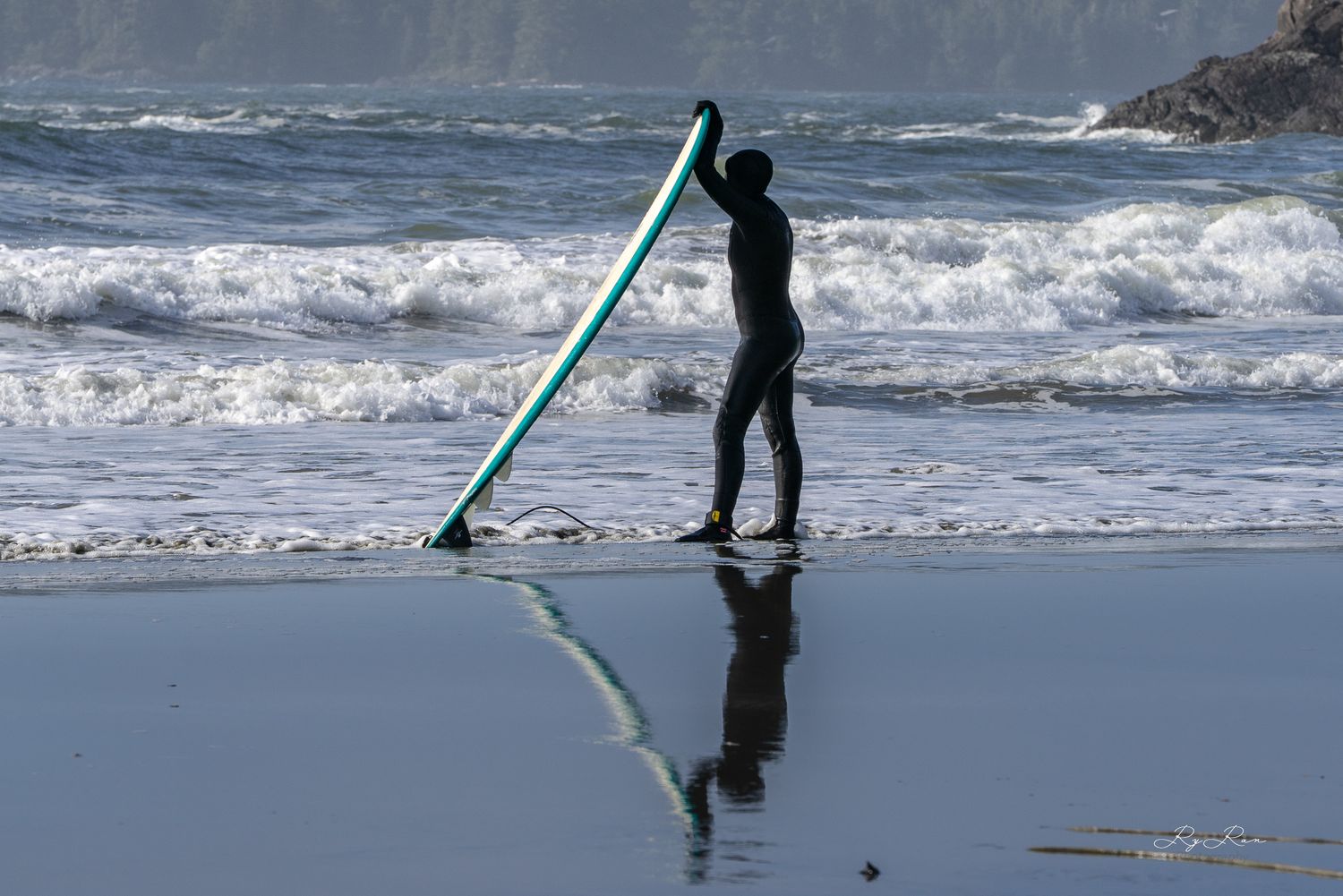 Surfer silhouetted against ocean waves carries long surfboard on reflective beach.