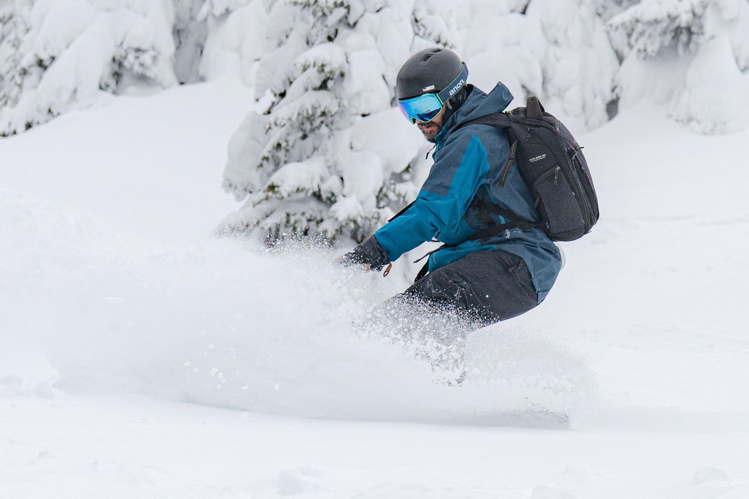 Snowboarder carving through fresh powder snow wearing blue gear and a backpack.
