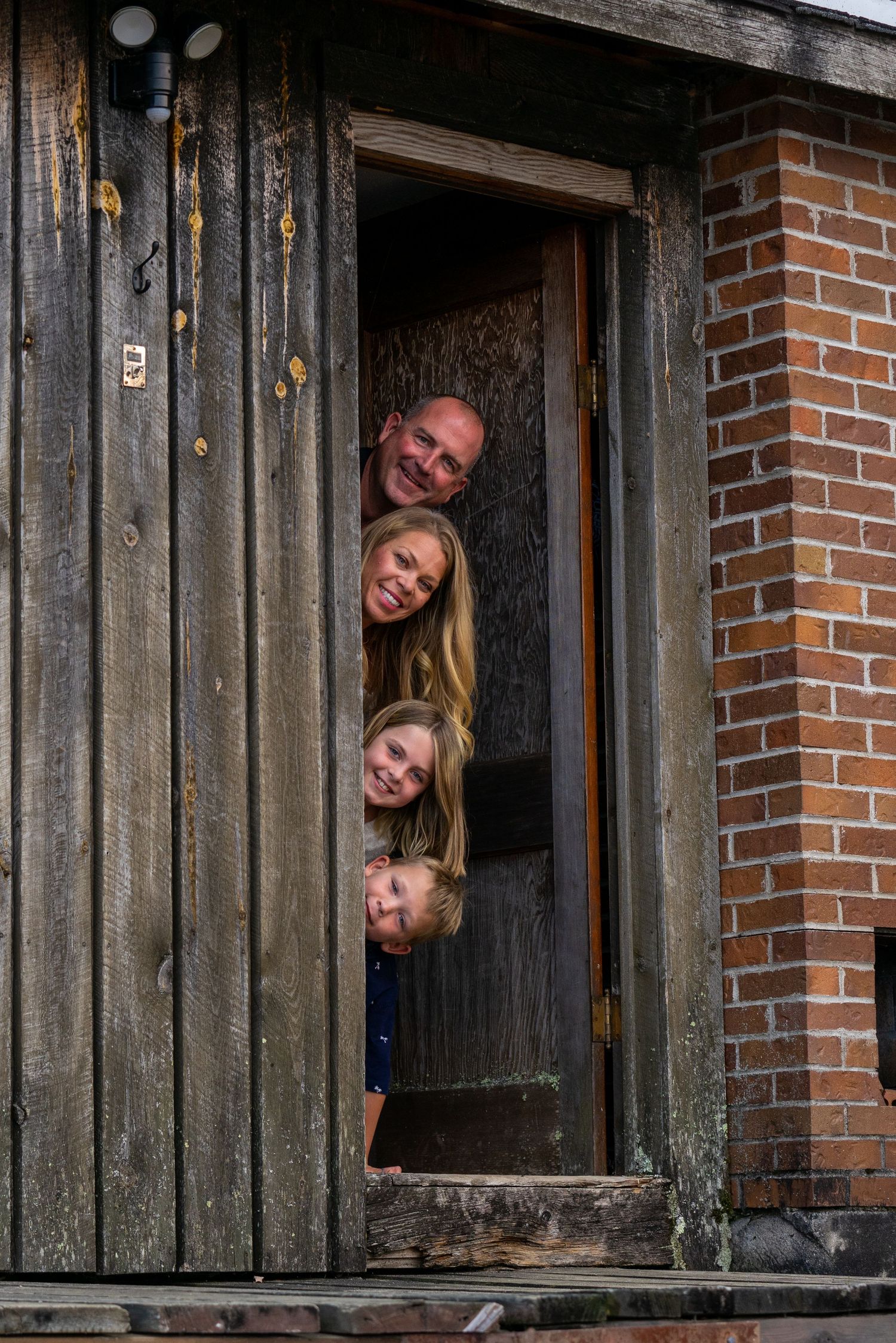 Family members playfully peek out from a rustic wooden doorway with brick accents.