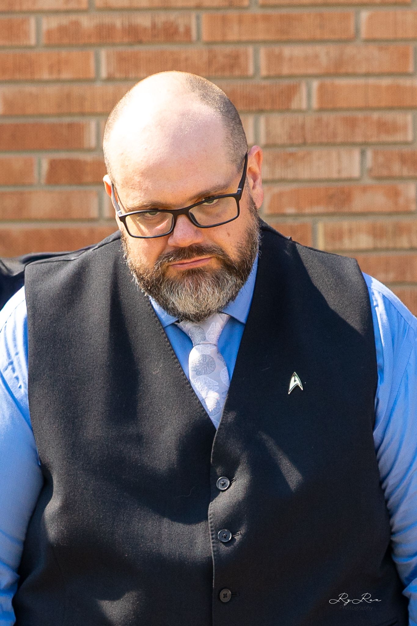 Portrait of person wearing glasses and blue tie with dark vest against brick wall background.