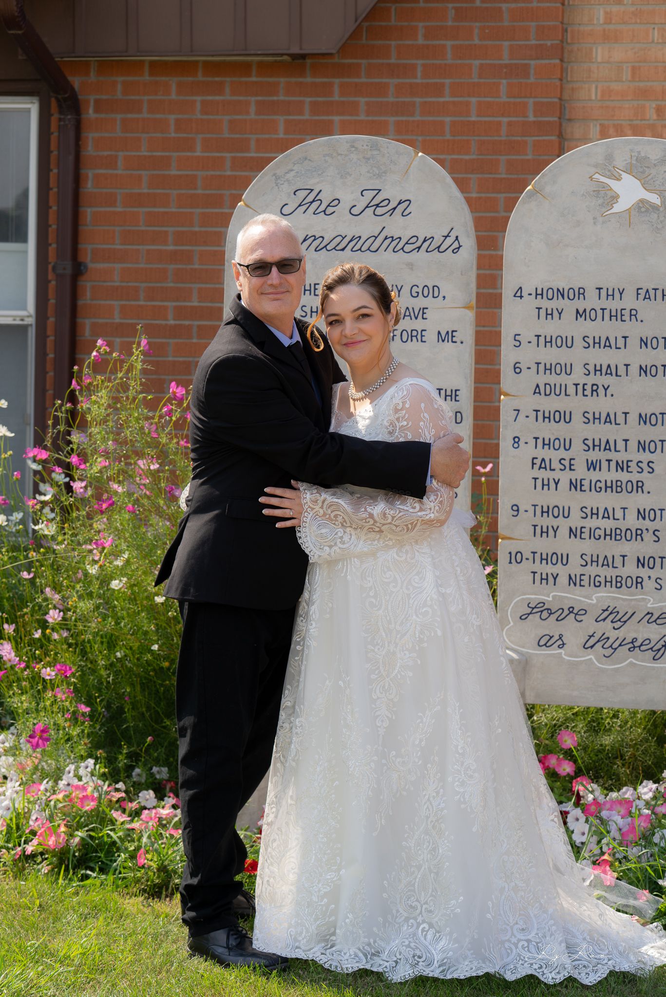 A wedding portrait in front of Ten Commandments stone tablets with pink flowers blooming in the garden.