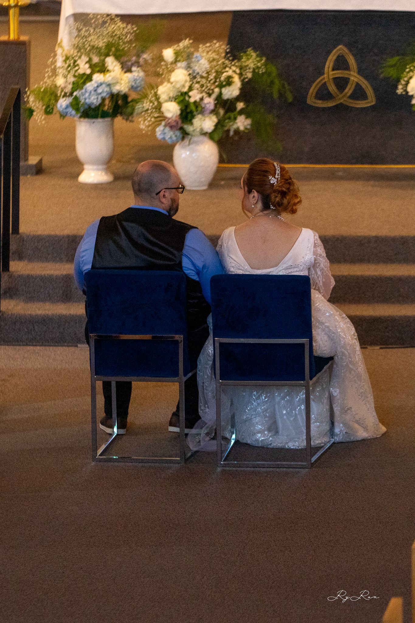 Couple seated at altar with floral arrangements and trinity symbol visible in background.