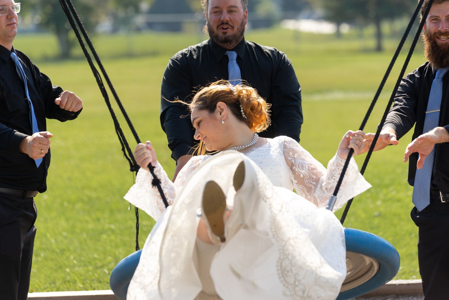 Wedding party members playing on a swing set in a sunny park setting.