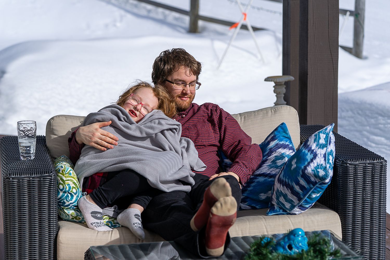 Two people share a cozy moment relaxing together on outdoor patio furniture with colorful blankets and pillows.