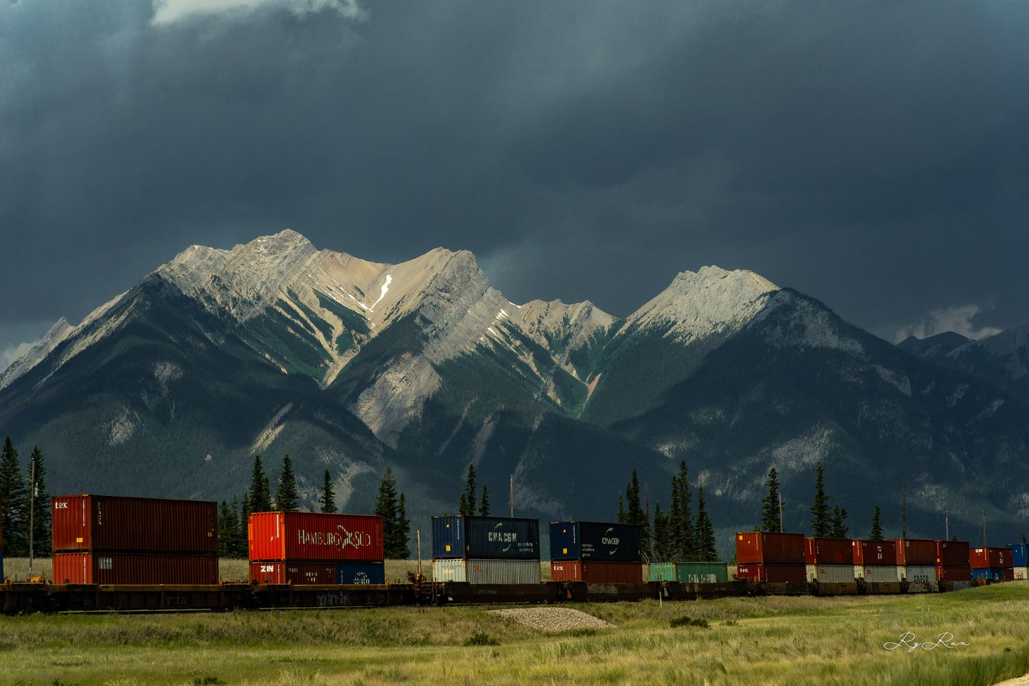 Freight train passes beneath dramatic storm clouds and snow-capped mountain peaks.