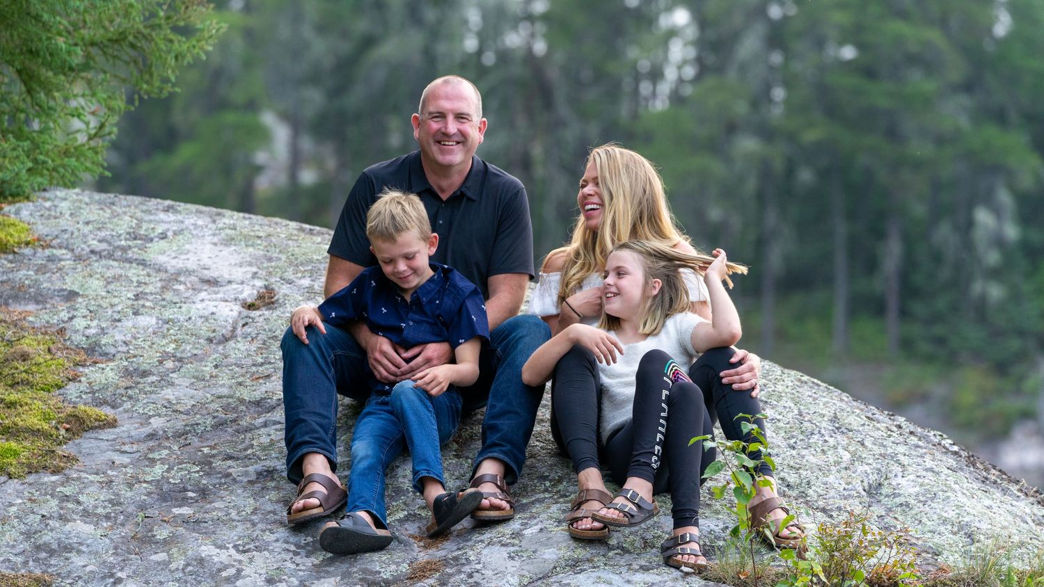 Family group sitting on large granite rock formation with evergreen forest backdrop.