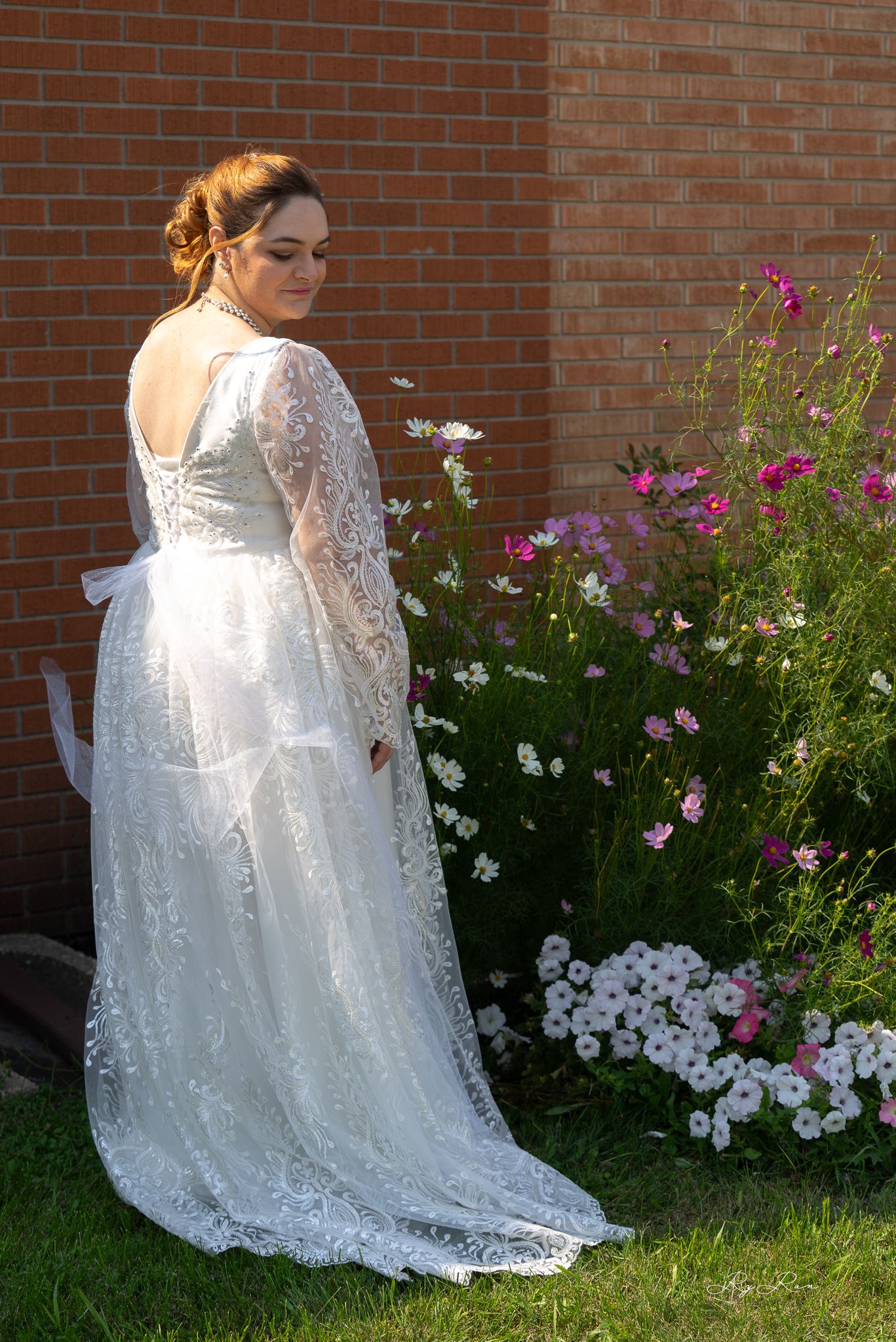 Beautiful lace wedding dress with long sleeves shown from behind against a brick wall with colorful garden flowers.
