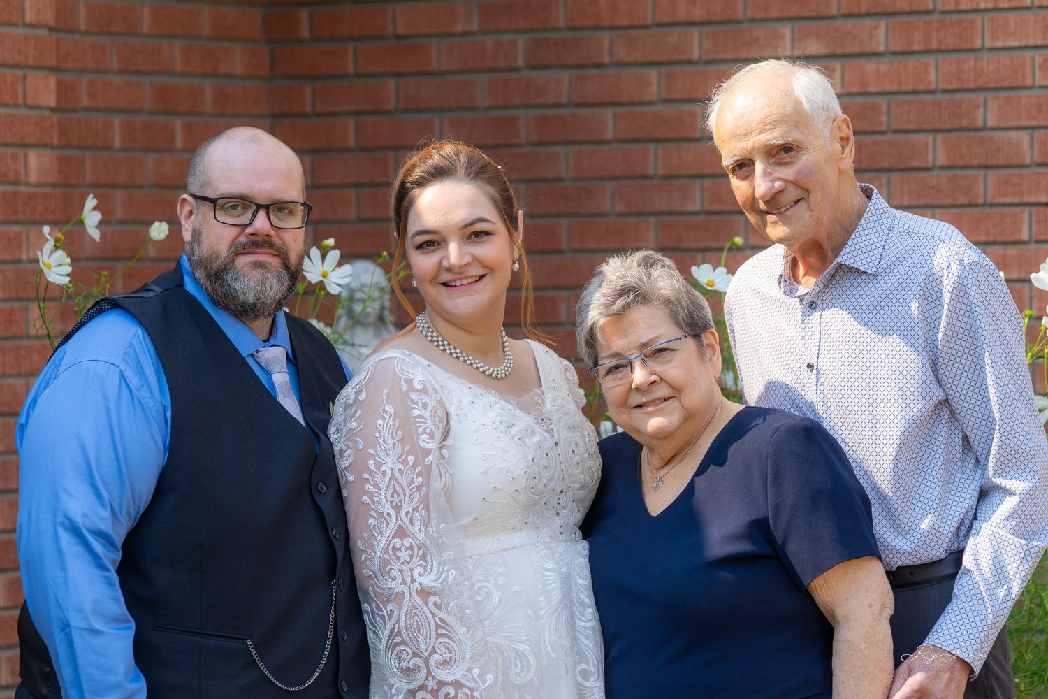 Smiling family group poses for wedding photo against brick wall with white flowers.