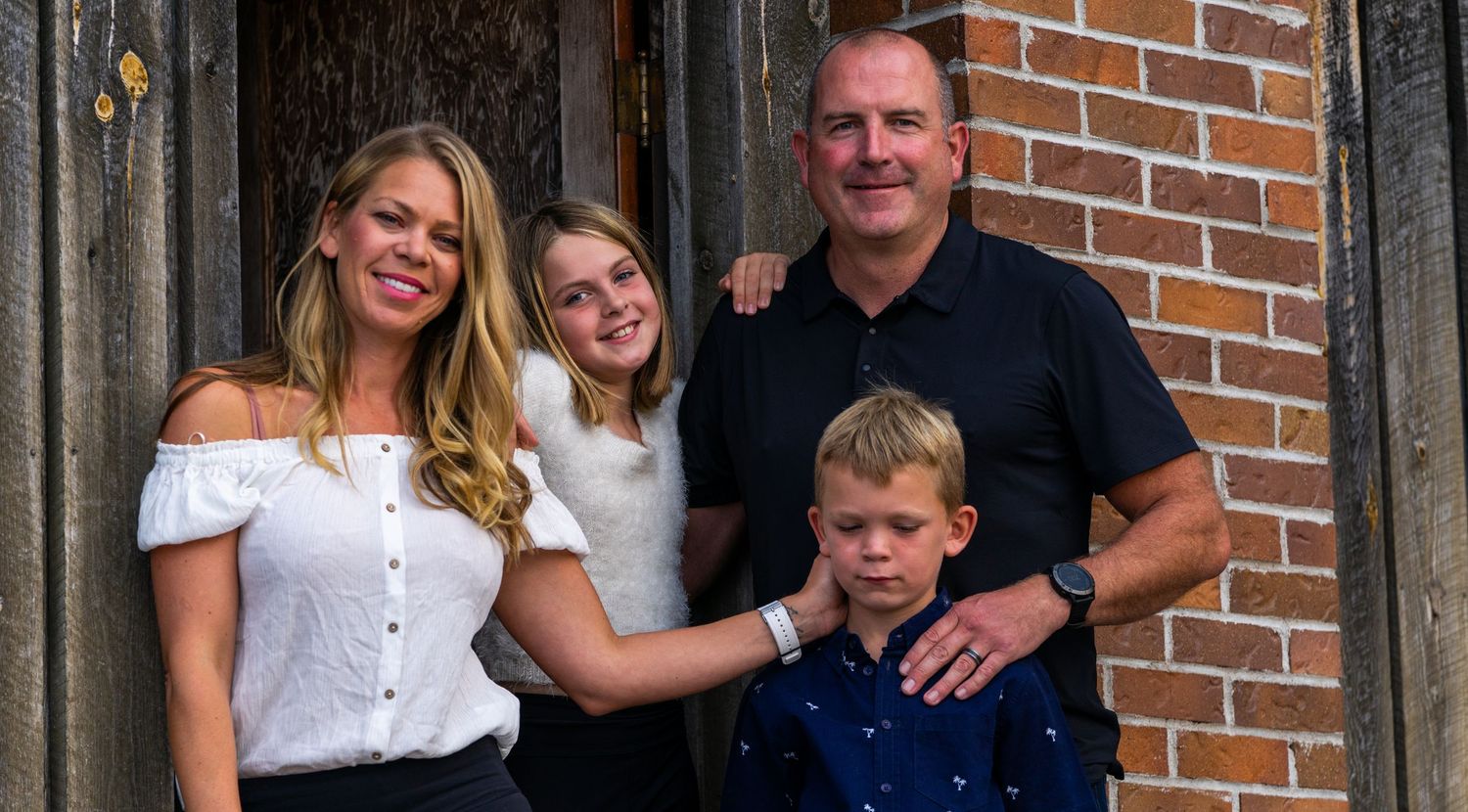 Casual family portrait taken in front of weathered wooden door with brick wall.