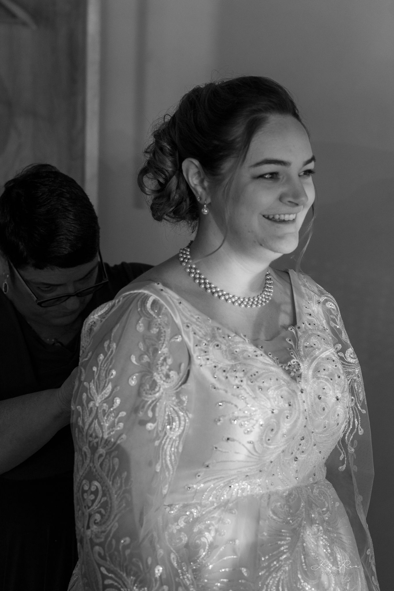 A bride in a white lace wedding dress with long sleeves and pearl necklace smiles during preparations.