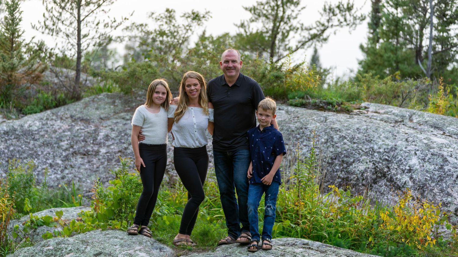 Family of four standing together on rocky terrain with evergreen trees in background.