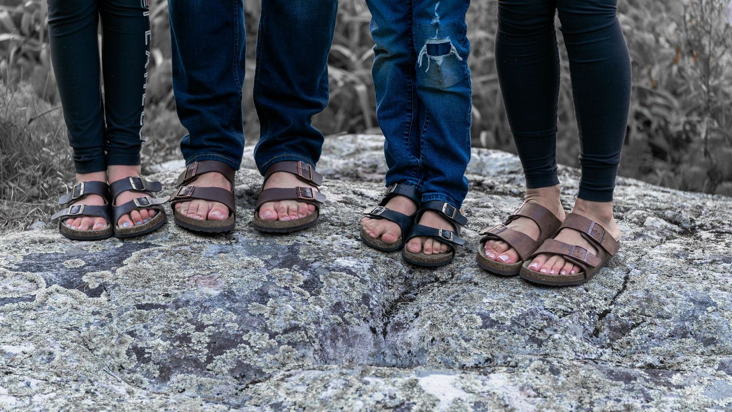 Close-up shot of four pairs of sandaled feet standing on rocky ground.