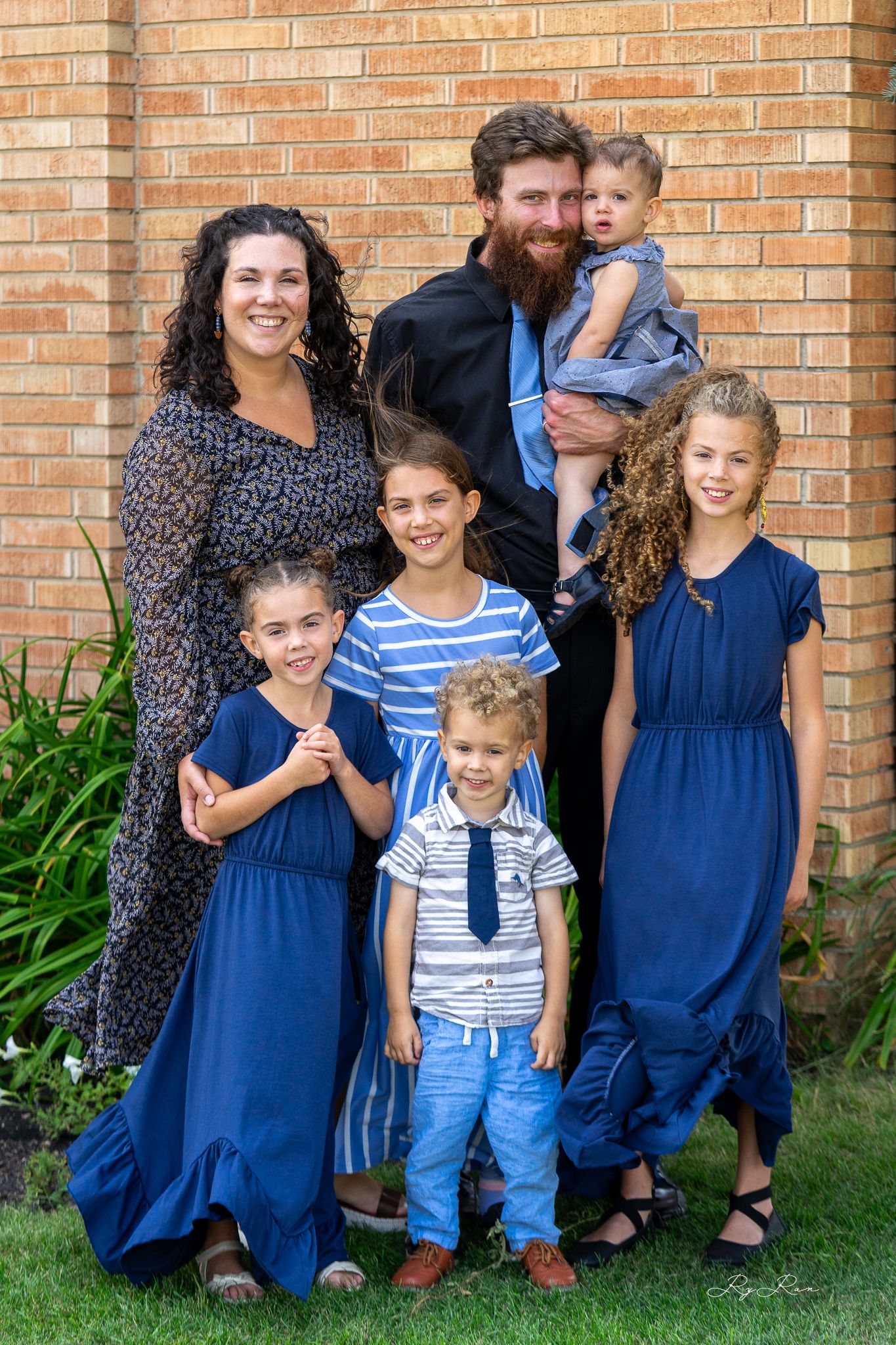 Family group portrait outdoors wearing coordinating blue outfits against brick wall.