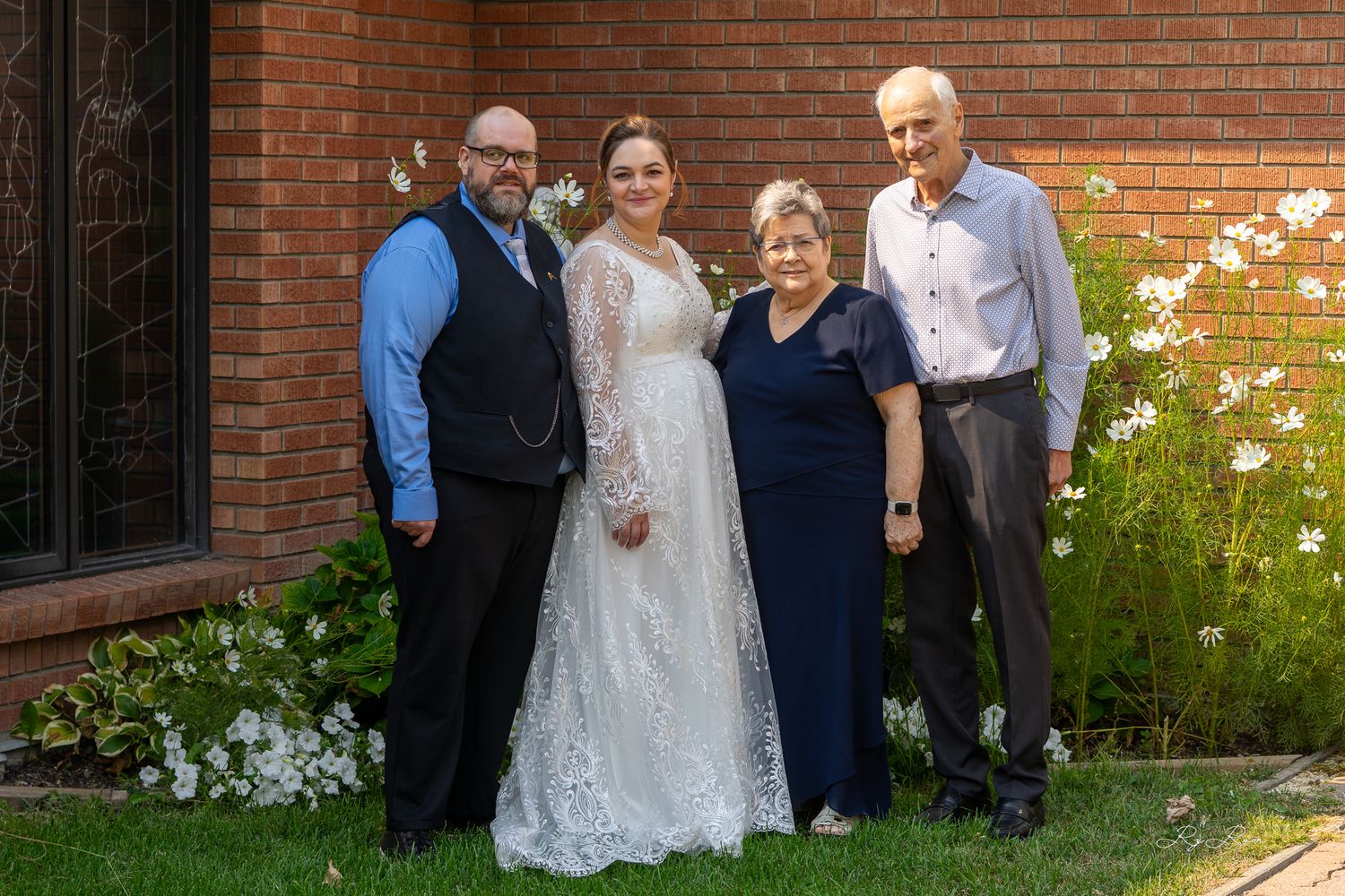 Family group stands together for formal wedding portrait by brick wall and garden.